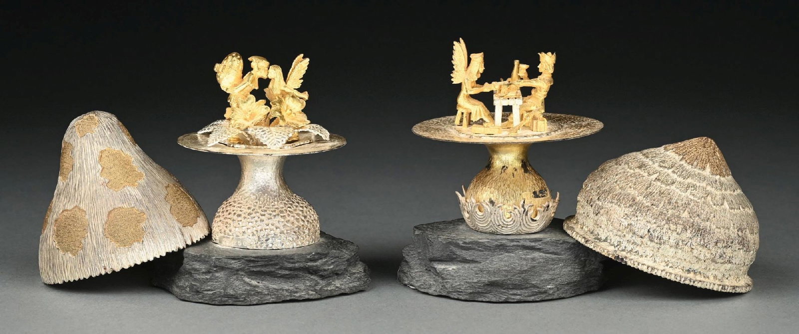 A PAIR OF SILVER MUSHROOMS ON SLATE BASES: Christopher Lawrence, London 1979, each with a textured cap lifting to a figural interior, one with a leprechaun kissing a fairy, the other with a King and Queen dining. Height: 4-5/8". Condition: Dim