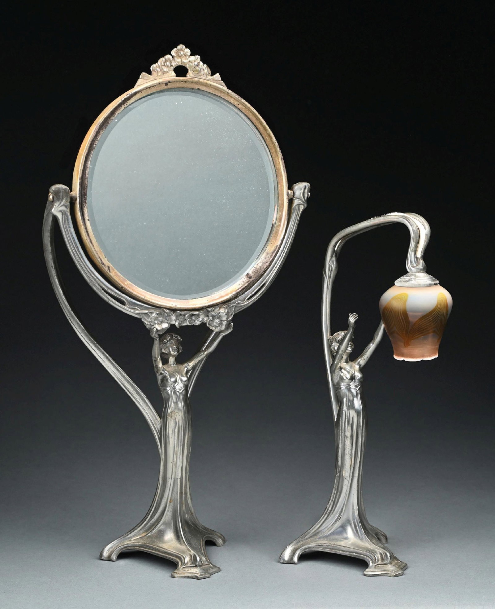 WMF ART NOUVEAU VANITY SUITE COMPROMISING A FIGURAL MIRROR AND BOUDOIR LAMP WITH TIFFANY SHADE, c.: A Wuttembergische Metalwarrenfabrik silverplated brass and glass suite. The Tiffany shade is etched M5302(c. 1900). The metalwork is unmarked, both the mirror and lamp are illustrated in Art Nouveau M