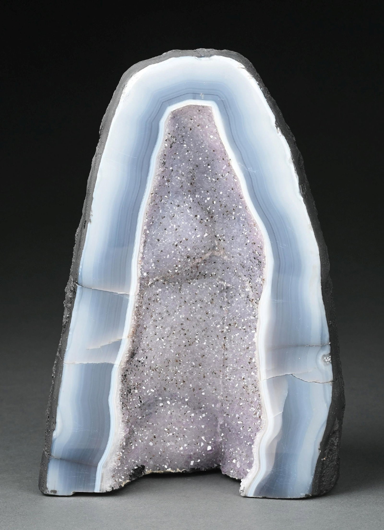 LARGE CATHEDRAL BOLIVIAN AMETHYST CRYSTAL GEODE WITH BLUE AGATE: A light lavender amethyst geode cathedral featuring a natural basalt outer shell with gray and blue agate banding. The interior displays light lavender druzy quartz with small black mineral inclusions