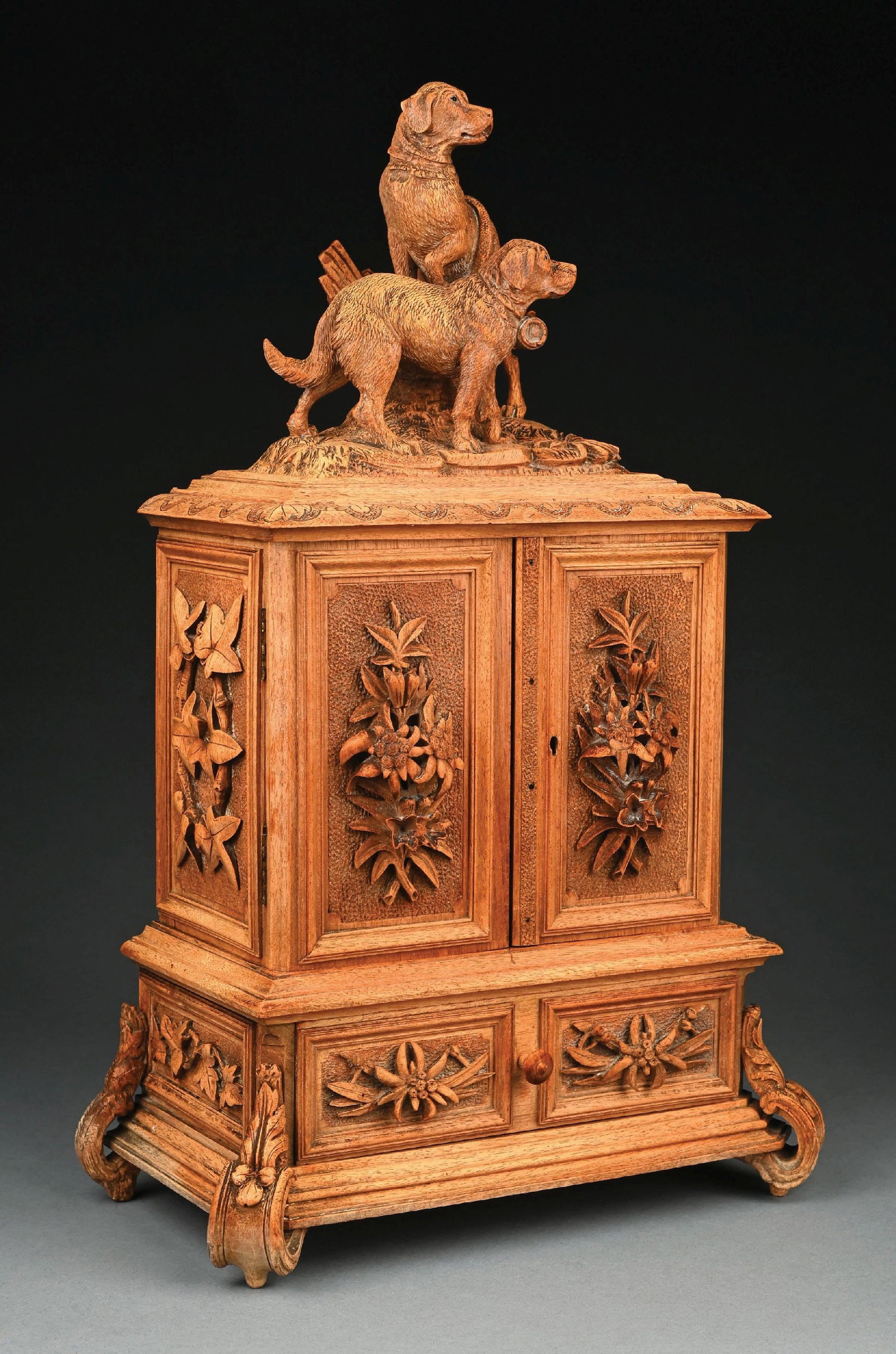 HAND-CARVED BLACK FOREST JEWELRY BOX: C. 1890–1920. A finely hand-carved Black Forest jewelry box featuring a sculptural group of hunting dogs mounted atop a stepped cabinet form. The box is richly decorated with deeply carved flora