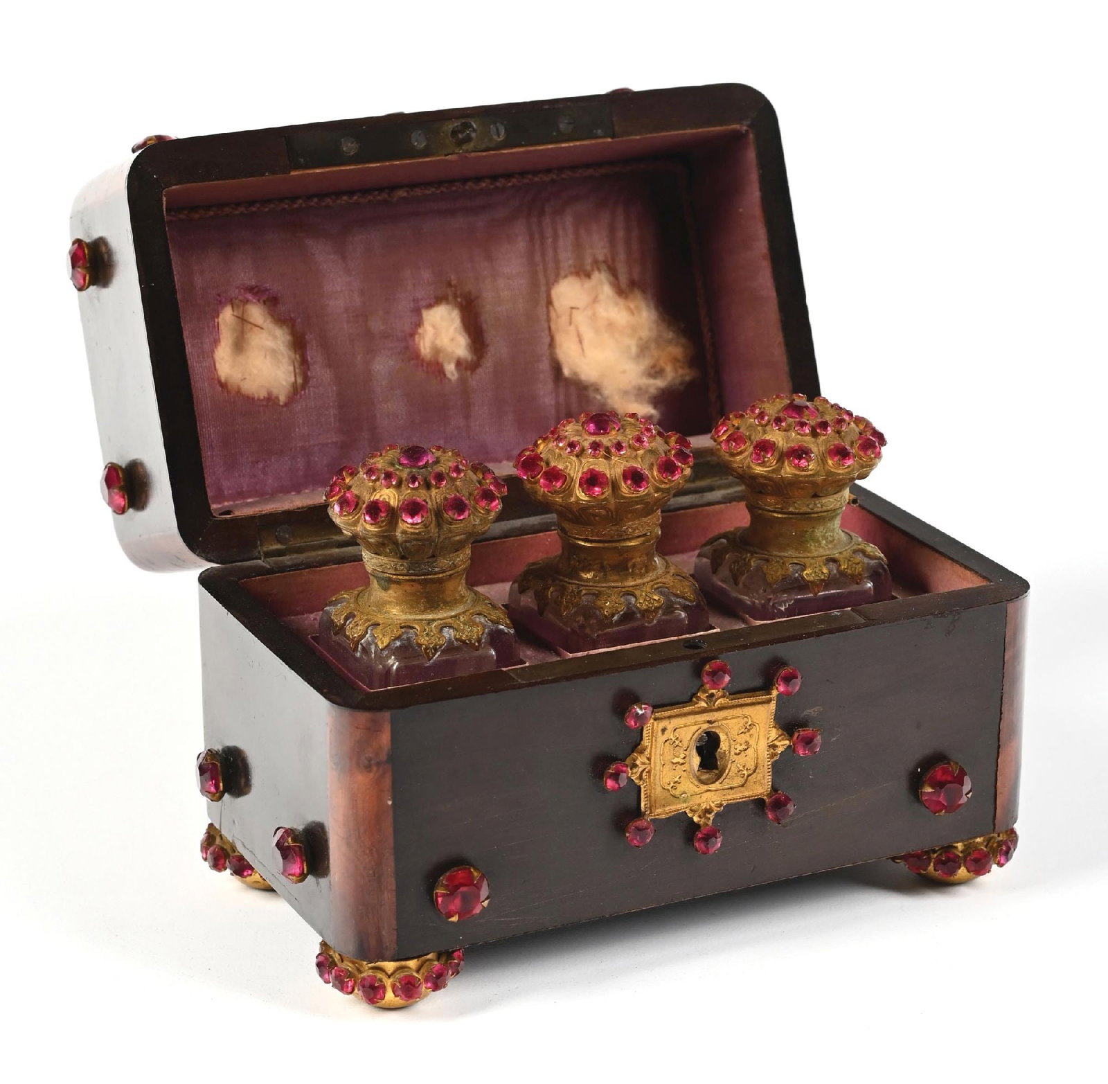PERFUME CASKET FEATURING THREE GLASS BOTTLES WITH ORNATE RHINESTONES: C. 1800s. An elaborate, deep rosewood perfume casket featuring red inlaid rhinestones into brass mounts. The casket features a decorative lock plate. The case interior is purple velvet. The case conta