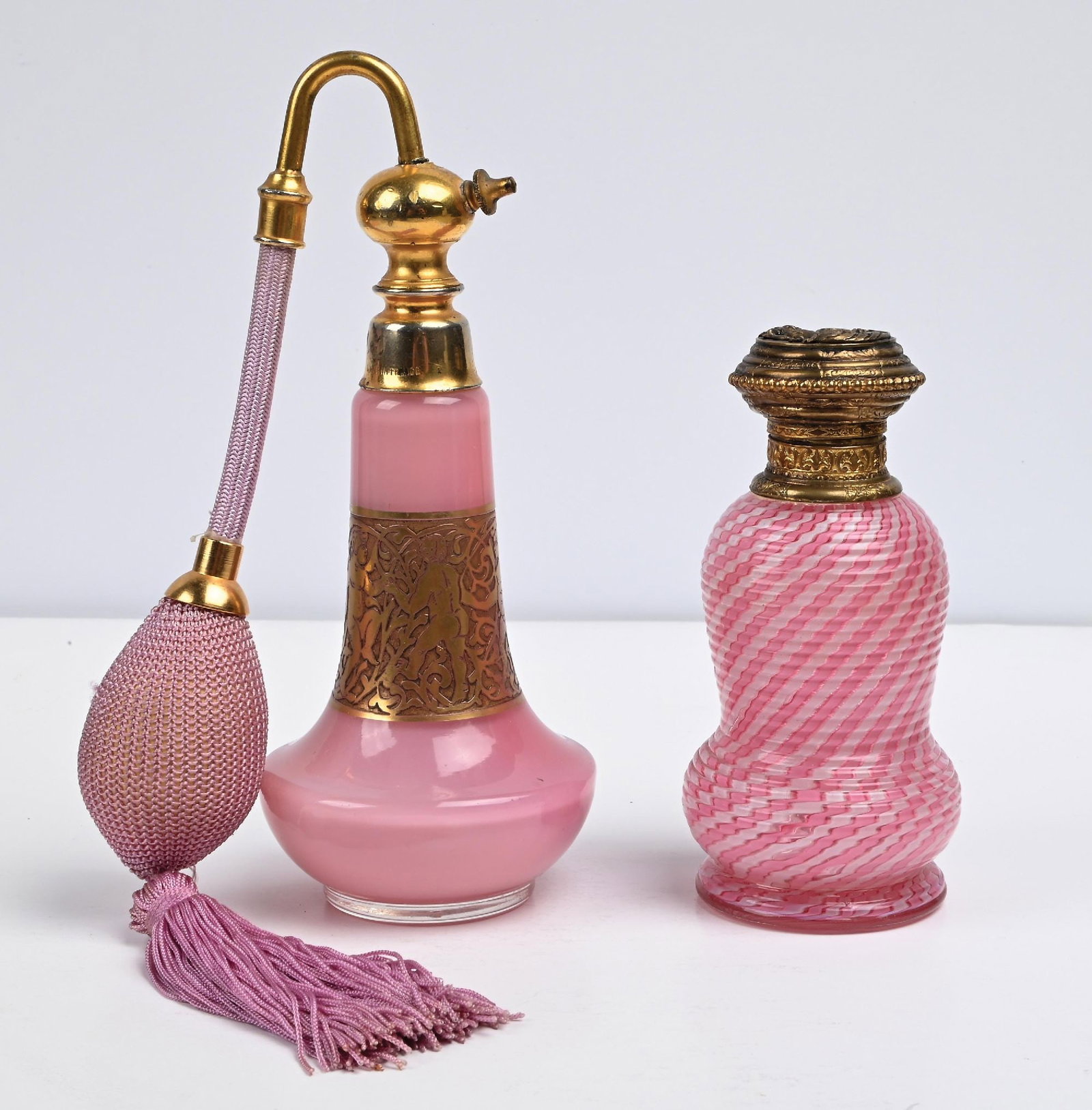 LOT OF 2: MOSER & LATTICINO PINK GLASS PERFUME BOTTLES: C. 1900s. (A) A lovely Art Deco opaque pink cased perfume atomizer featuring a gilt gold-tone figural overlay around the neck. The perfume includes an attached pink woven mesh squeeze puff with tassel
