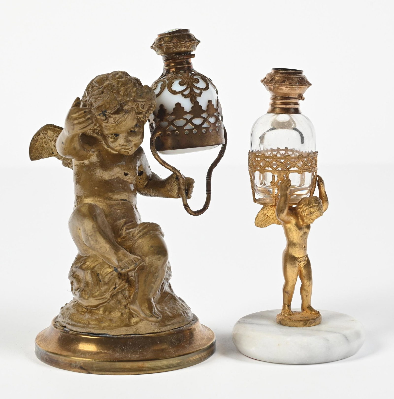 LOT OF 2: CHERUB PERFUME HOLDERS: C. 1800s. (A) A gorgeous gold-toned metal cherub perfume holder with a white opaline glass perfume and porcelain cap. Light surface wear throughout, as expected with age. (B) A smaller brass perfume h