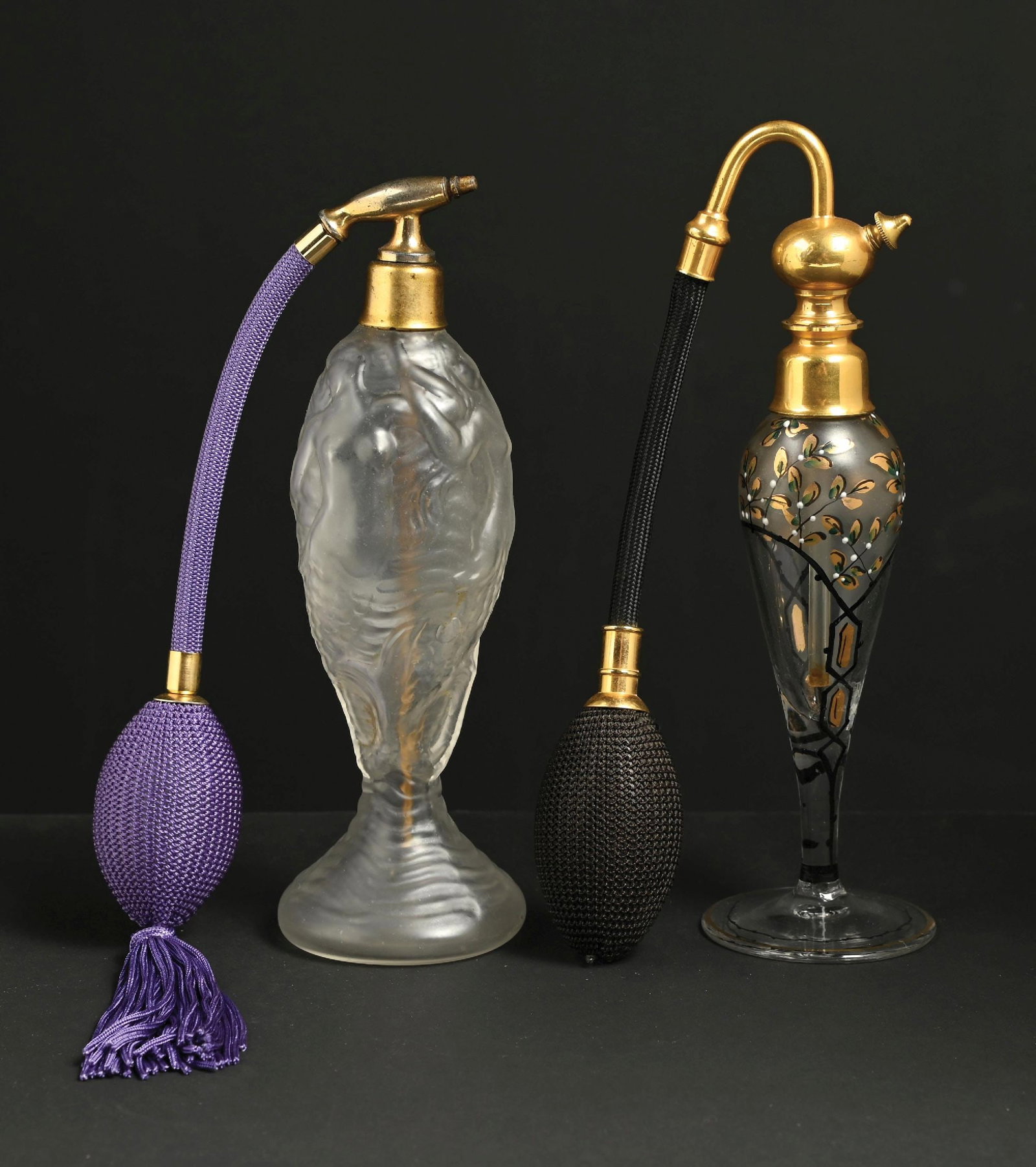 LOT OF 2: VINTAGE GLASS PERFUMES WITH ATOMIZERS: C. 1900s. (A) An attractive frosted glass perfume atomizer featuring three mermaids. The perfume has gold-toned hardware. The perfume has a teardrop shape and a stem, with a purple hose and a function