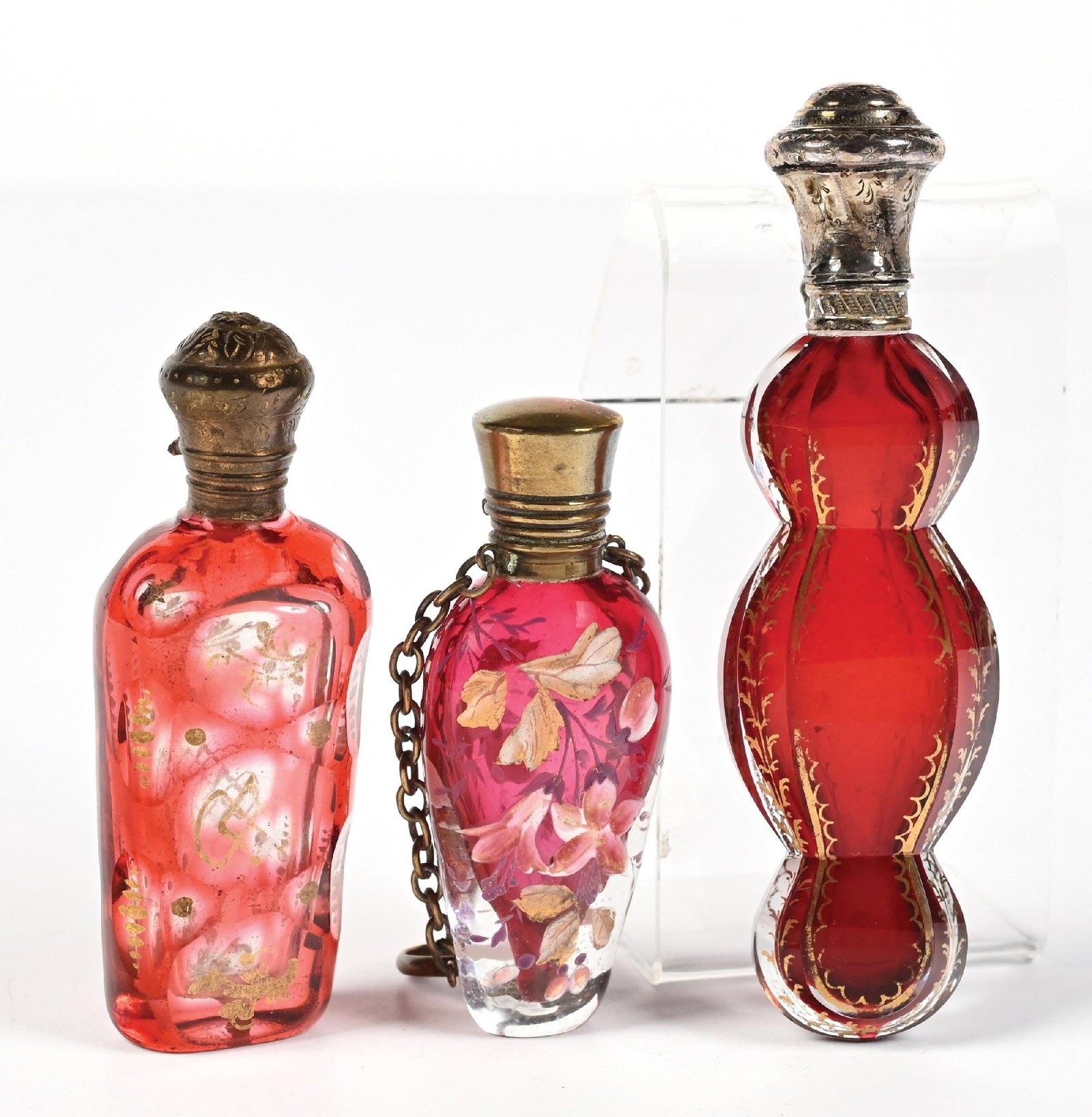 LOT OF 3: RUBY RED GLASS PERFUME BOTTLES: C. 1800s. A collection of three red ruby glass perfume bottles, including a cut-to-clear and a French cased glass, with ornate gold-tone caps and floral enamel work, all in great condition. Largest: 1