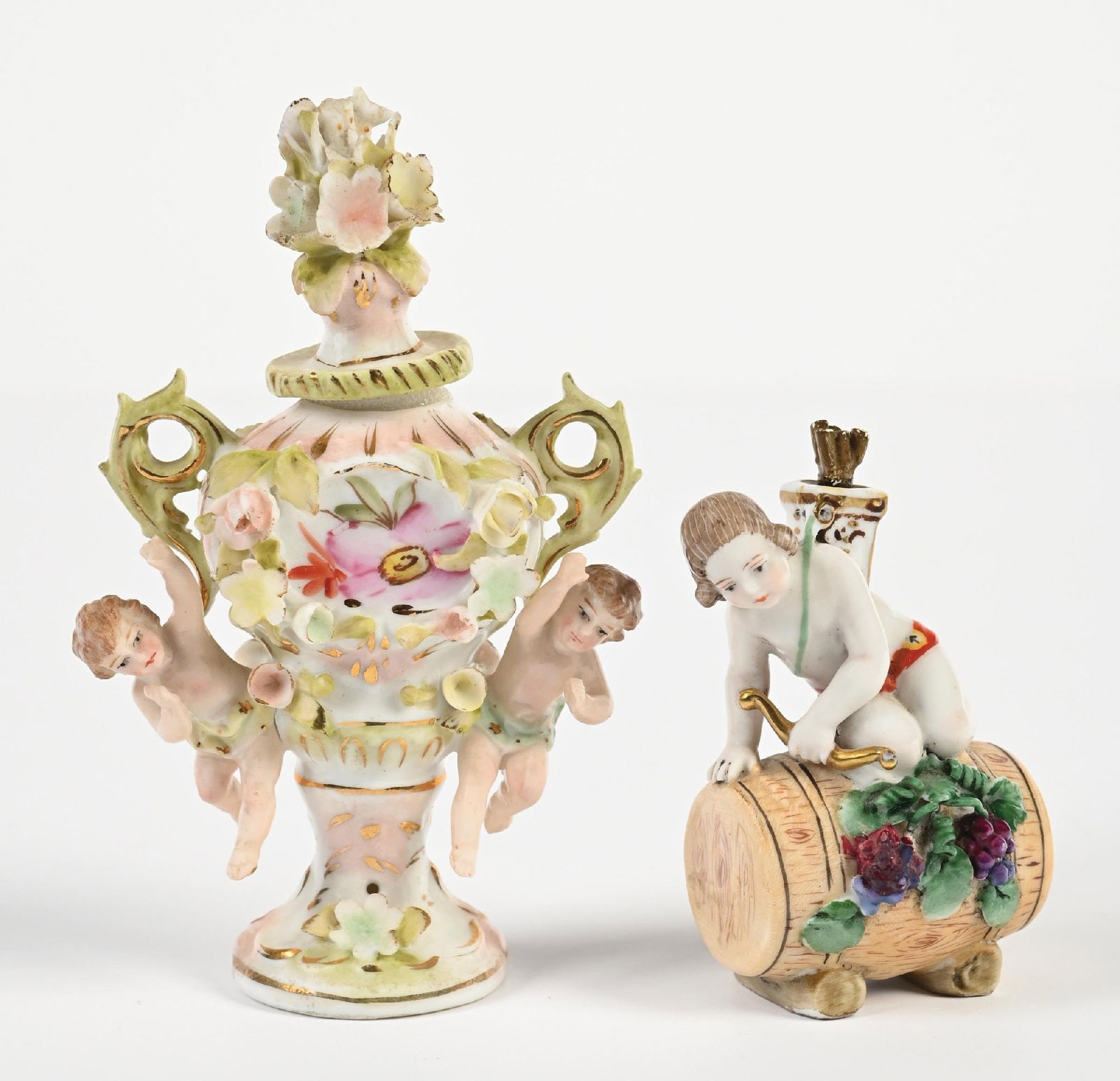 LOT OF 2: SITZENDORF FIGURAL PORCELAIN PERFUME BOTTLES: C.1900–1925. Two Sitzendorf porcelain figural perfume bottles each modeled with applied putti and floral encrustation forming the vessel body. One example features a cherub seated beside a barre