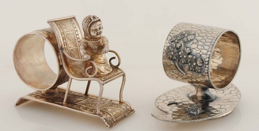 Lot of 2: Figural Napkin Rings.: First napkin ring is a Kate Greenaway-type baby seated on a chair. The napkin holder, base, and chair are highly decorated with a plethora of Victorian-era ornamentation and flowing lines. Marked 'Mid