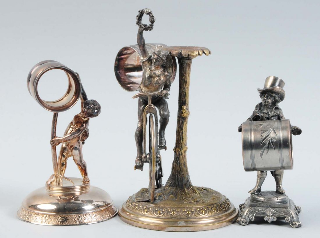Lot of 3: Figural Napkin Rings.: The lot includes a napkin ring that depicts a tall, draped cherub atop a domed round base who is balancing a napkin holder atop a tree branch. It is marked "Simpson, Hall, Miller, & Co., No. 102". It