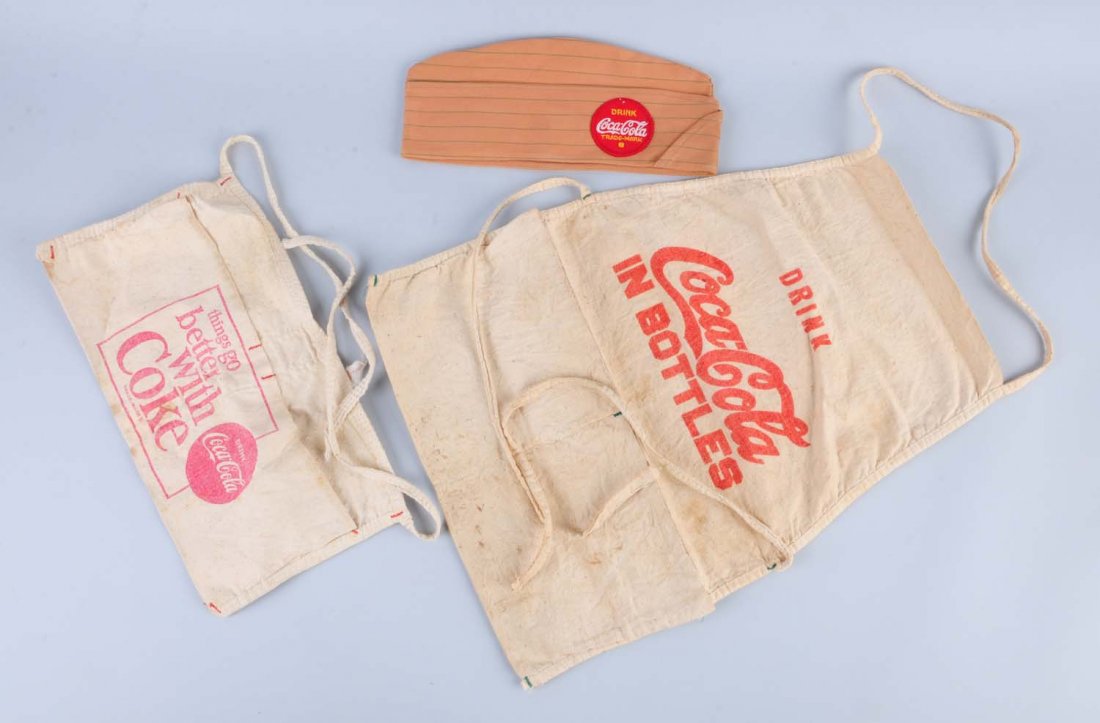 Lot of Coca - Cola Aprons and Cap.: This lot includes two Coca - Cola aprons with overall wear and cap that appears not to have been used. Condition (Very Good - Excellent). Size Largest: 28" L.