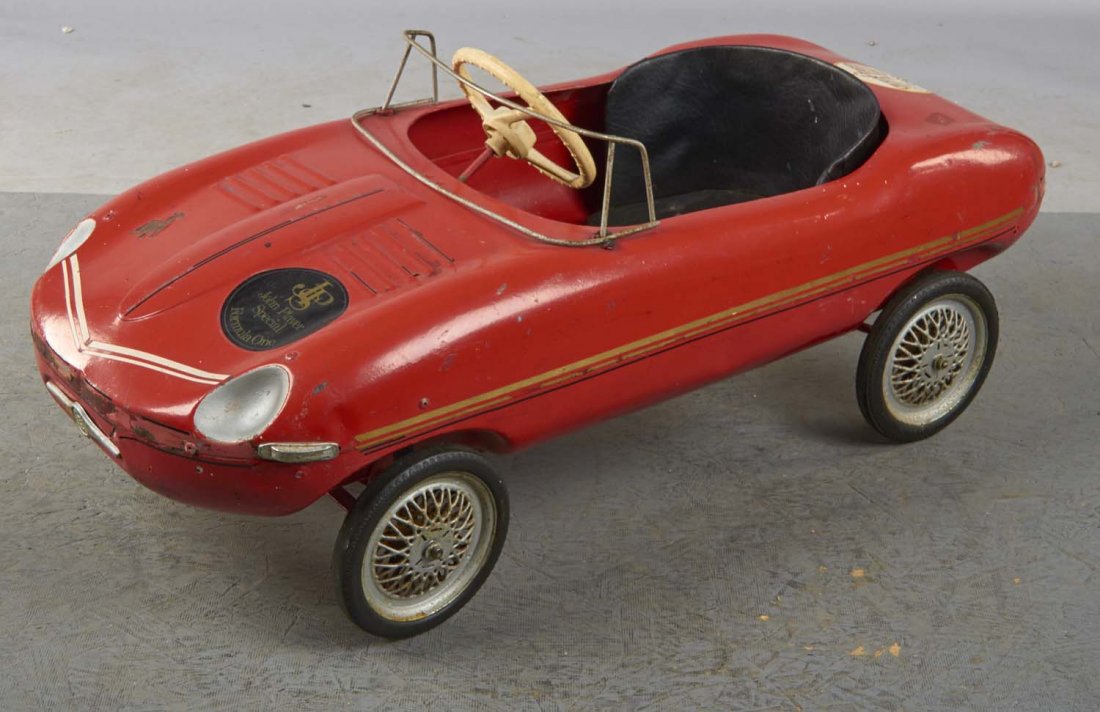 Pressed Steel Triang E-Type Jaguar Pedal Car: This red pedal car with black leather upholstered seat features white plastic steering wheel, spoke wheels, and decals for Dunlop and John Player Special Formula One. Condition (Good). Size 40" x 18" 