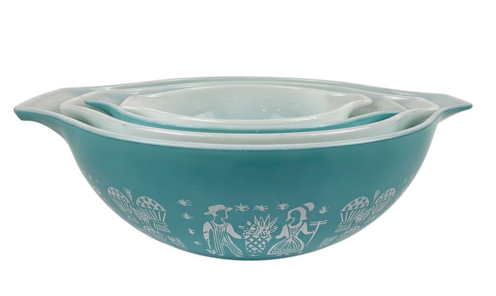Pyrex Amish Butterprint Turquoise and White Bowl Set: Pyrex mixing bowls featuring the Butterprint pattern in turquoise and white. The patterned bowls depict a scene of a farmer and woman holding produce, surrounded by farm motifs. Nesting design with tw