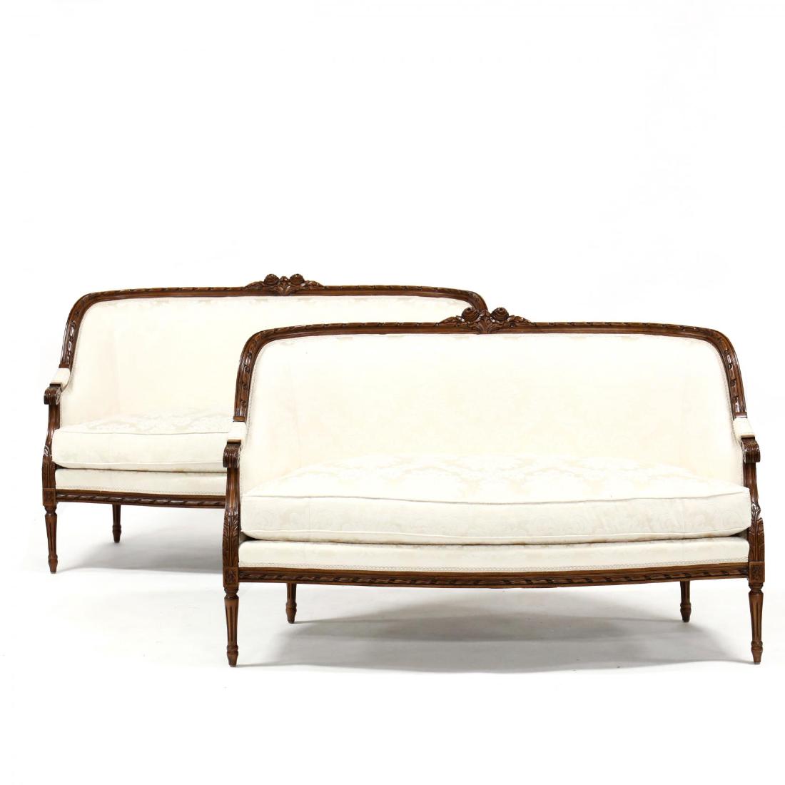 Pair of Louis XVI Style Carved Walnut Settees: 20th century, relief carved floral crest rail, acanthus carved arms, on fluted legs, silk floral damask upholstery with loose cushion. 37 x 60 x 34 in. From the Collection of Mr. & Mrs. McDara Folan,