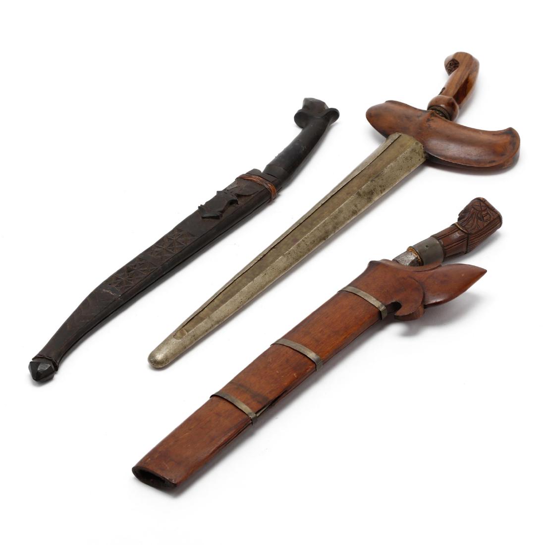 Three Vintage Philippine Knives: early 20th century, to include a kris with metal scabbard (19 in.); a bolo knife with wooden scabbard; a bolo with wooden scabbard carved with a relief shield representing the U.S. administration. Ad