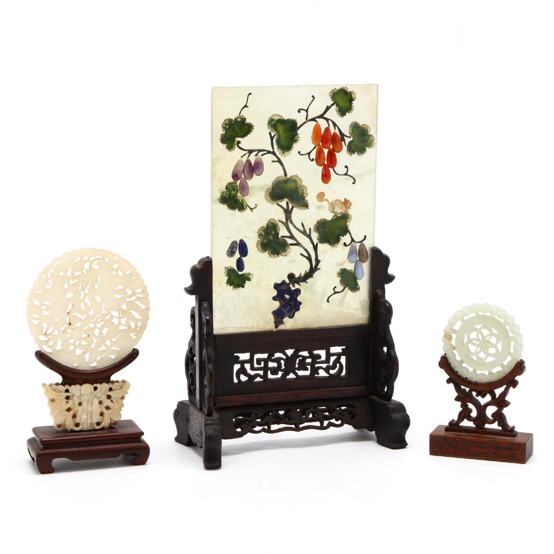 Three Chinese Jade Carvings in Stands: Three Chinese Jade Carvings in Stands includes a vertical jade panel embellished with semi-precious stones mounted in a carved wooden table screen mount, a medium carved jade disc with figures and Bud