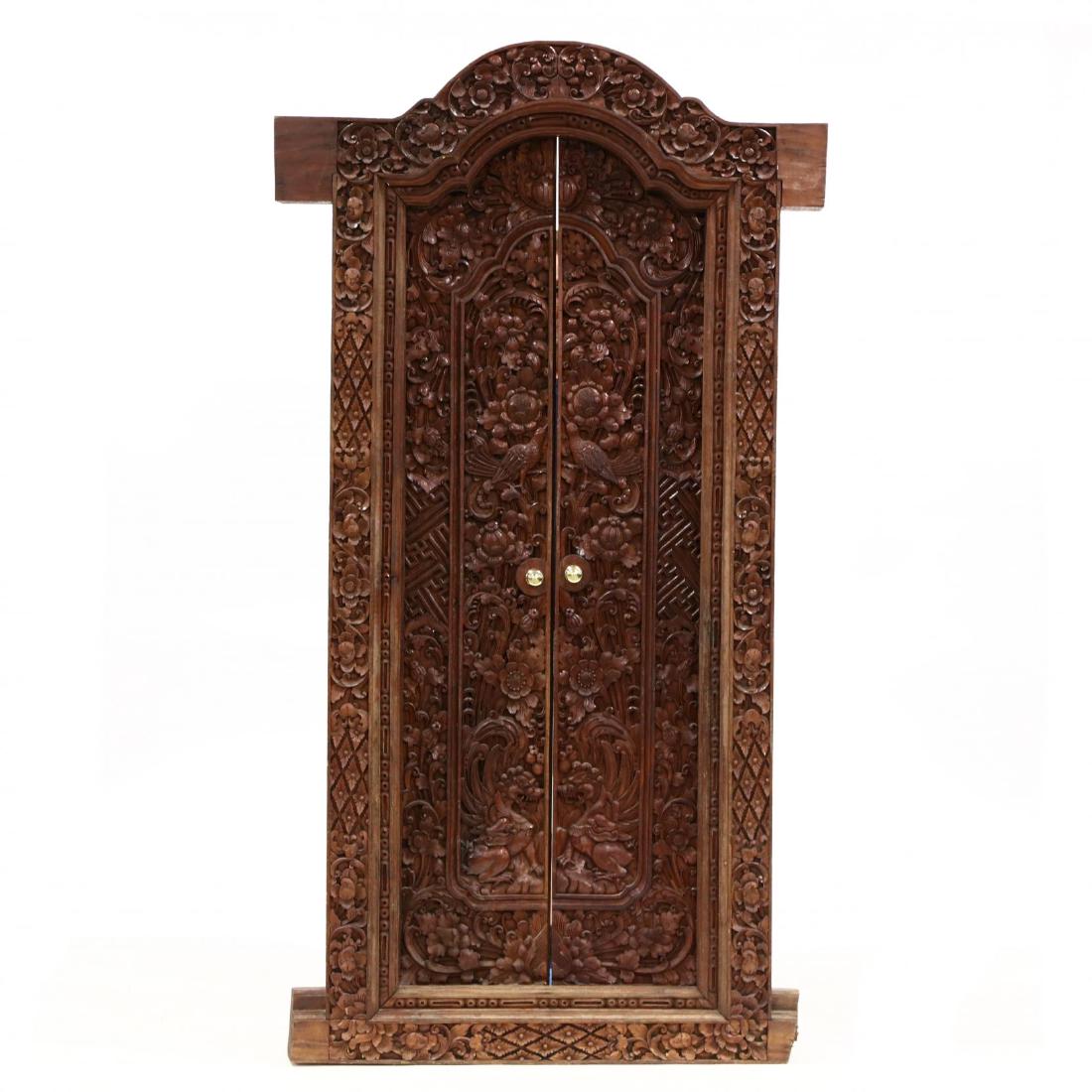 A Balinese Relief Carved Door Frame with Doors: A Balinese Relief Carved Door Frame with Doorslate 20th century, mahogany, thick carved posts, arched surround with flowers and diapered panels having inset peg hinged doors with repeating floral carv