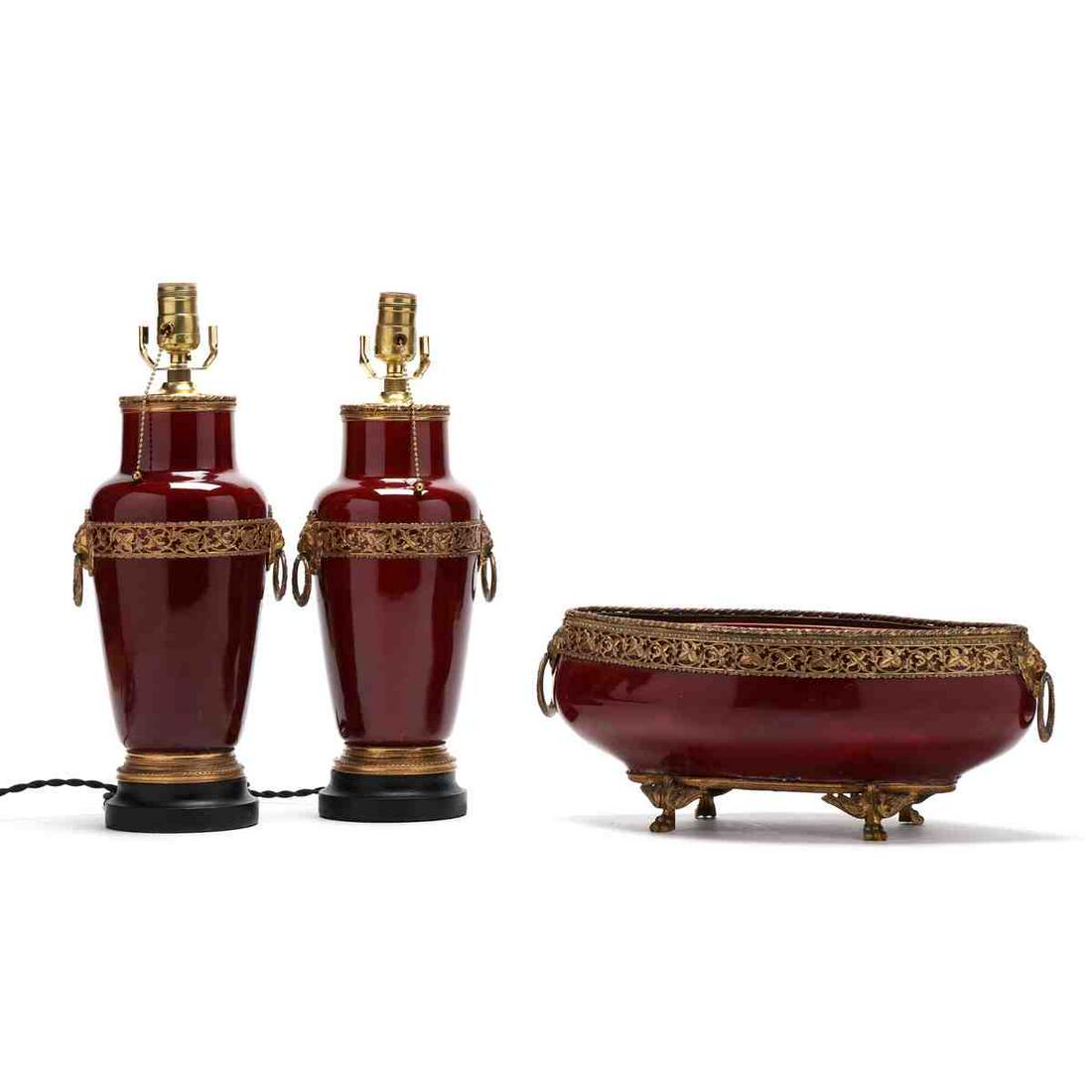 French Ormolu Mounted Oxblood Porcelain Lamp Garniture: French Ormolu Mounted Oxblood Porcelain Lamp Garniture early 20th century, including a pair of jar form lamps mounted to ebonized wood bases and a lozenge form pedestal bowl, all with lion mask ring h