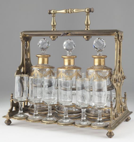 Baccarat Crystal Decanter Set in Tantalus: Baccarat Crystal Decanter Set in Tantalus includes (3) cut crystal decanters (8 in.) with gilt foliate swags and Greek key design, faceted stoppers, and (12) stems (4.25 in.) within locking brass cadd