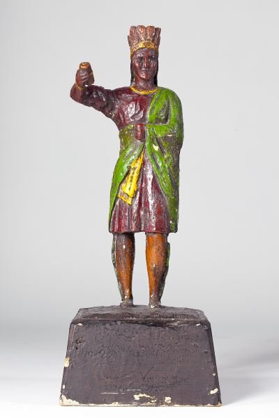 Polychrome Painted Counter-Top Cigar Store Indian: Polychrome Painted Counter-Top Cigar Store Indianlate 19th century, carved pine with allover gesso and paint, the figure wears a feathered headdress, cloak and leggings while holding aloft a handful o
