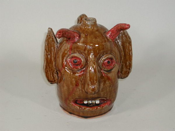Reggie Meaders Devil Face Jug,: Reggie Meaders Devil Face Jug, brown glaze w/red highlights to eyes, horns and mouth, clay teeth, printed ears, inscribed "Reggie Meaders" on head, one horn w/small chip, 9.5"h