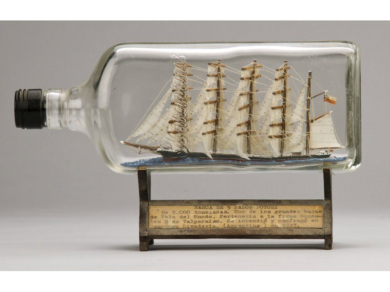 Antique 1923 Glass Five Masted Ship in a Bottle: Glass Ship in a Bottle of Five Masted Sailing Ship. The glass ship in an old Ballentine Whiskey bottle with wooden stand titled in Spanish which details the specifications of the Barca de V Palos Poto