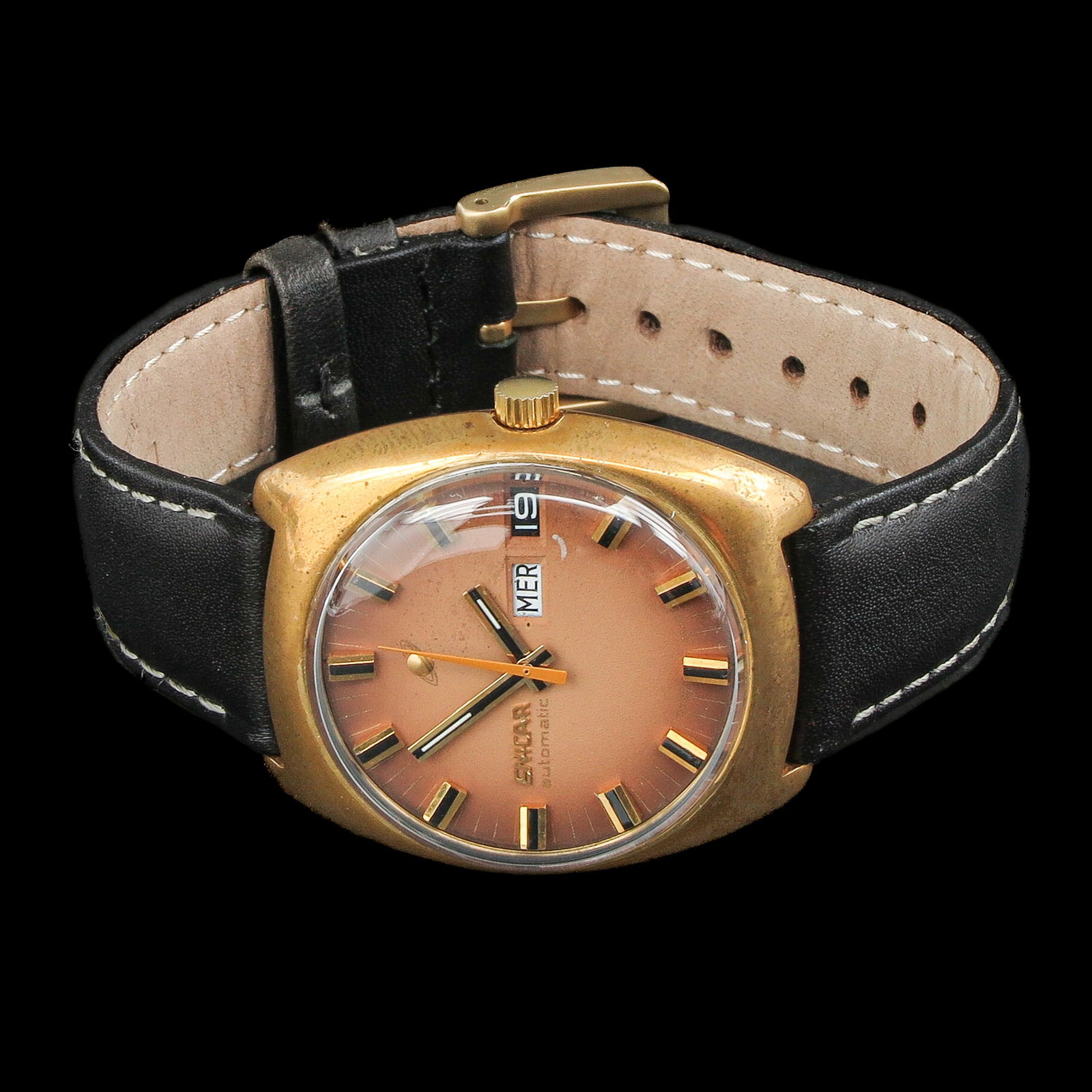 A Mens Watch: Enicar, with a gold-coloured cushion-shaped case, copper sunray dial with black and gold-coloured indexes and a date display at 3 o’clock. Equipped with a black leather strap with white stitching, c