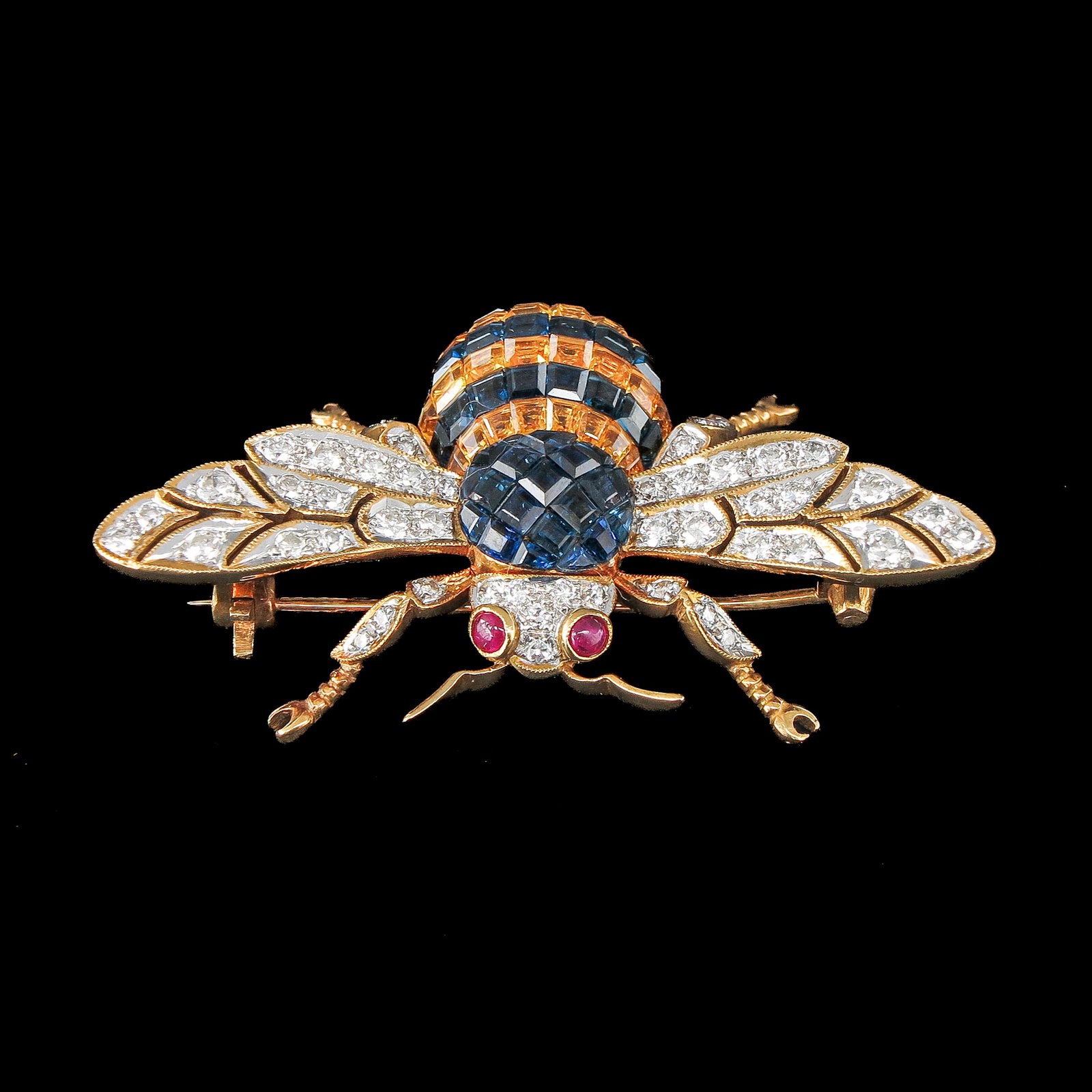 A Fine 18KG Jeweled Brooch: 18 Krt. gold, from the 1950s, in the shape of a bee, set with square-cut sapphires and diamonds, the 2 loops are cabochon-cut rubies, 50 x 30 mm