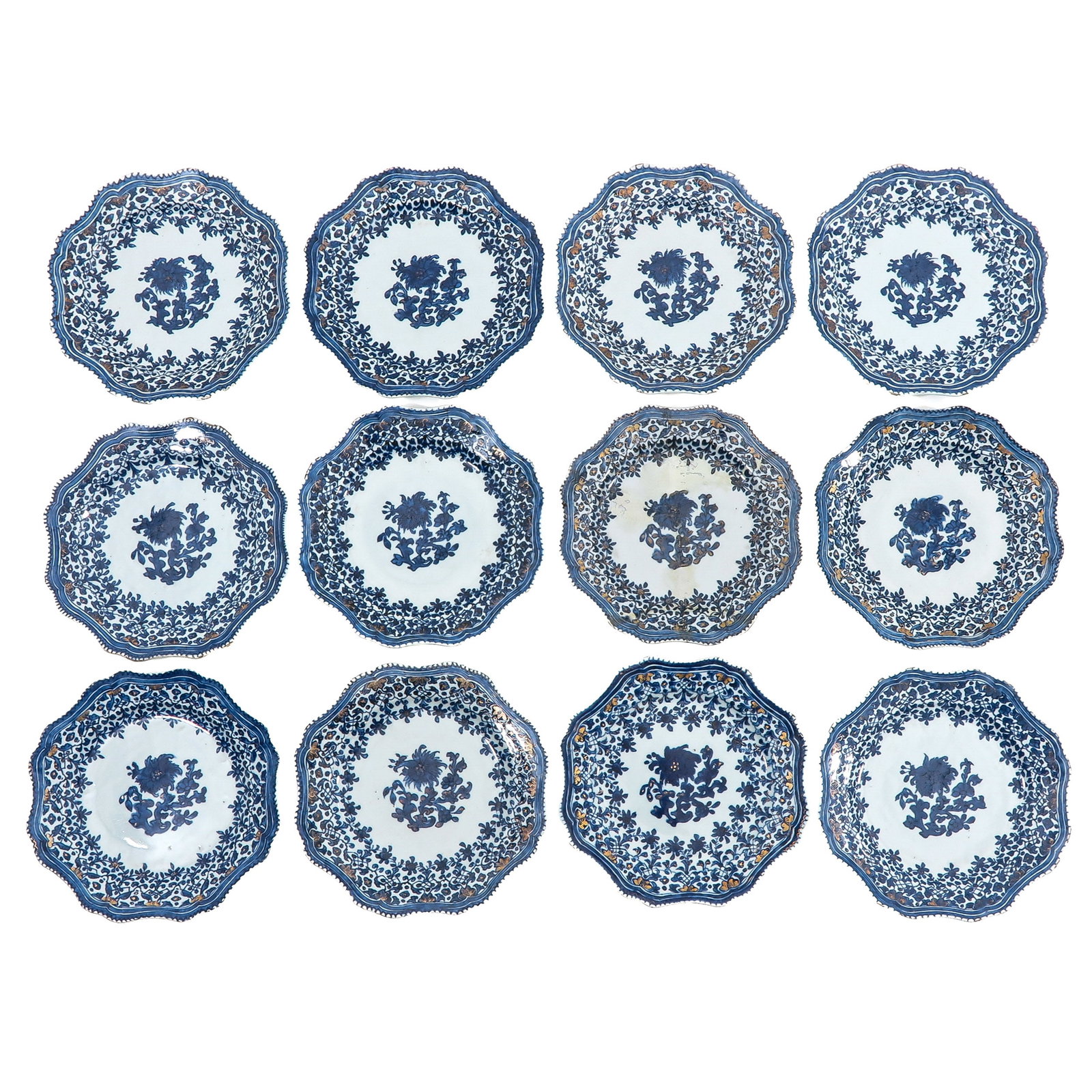 A Series of 12 Blue and White Plates: Scalloped rims, decorated with central floral sprays within dense foliate borders with gilt accents, Qianlong Period, 23 cm in diameter, chip and hairline.