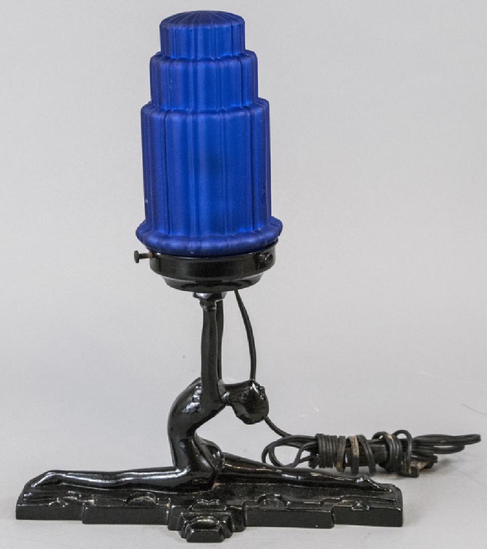 Art Deco Female Nude Figural Table Lamp: Art Deco Female Nude Figural Table Lamp. This table lamp having an Art Deco style features a figural female nude as its base supporting the upper light. Includes blue glass shade with tiered geometric