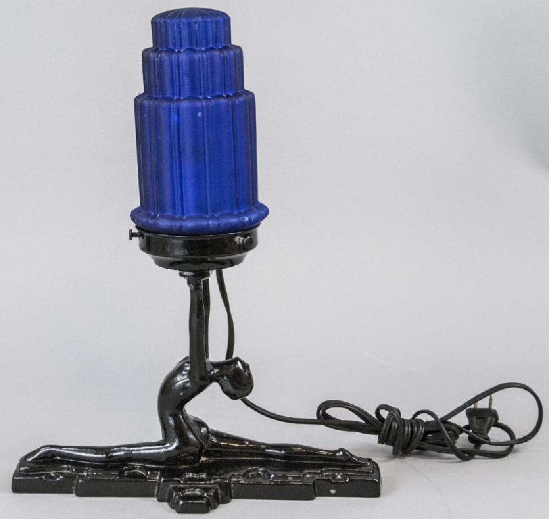 Art Deco Female Nude Figural Table Lamp: Art Deco Female Nude Figural Table Lamp. This table lamp having an Art Deco style features a figural female nude as its base supporting the upper light. Includes blue glass shade with tiered geometric