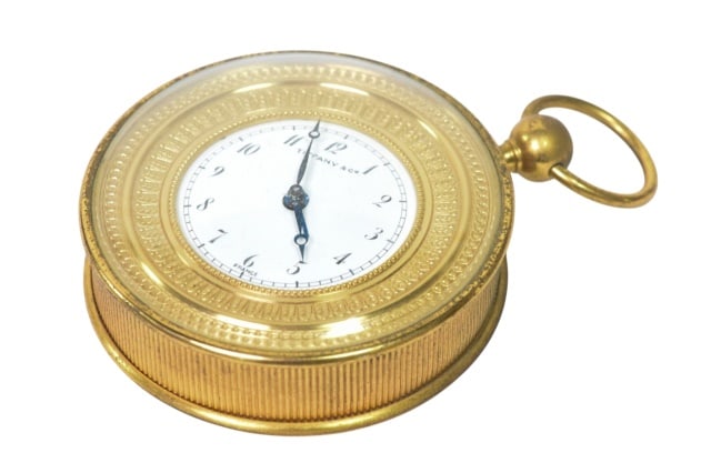 Tiffany & Co. Round Gold Tone Travel Alarm Clock: Vintage Tiffany & Co. Round Brass and Gold Tone Travel Alarm Clock with White Enamel Dial Black arabic Numerals and a Folding Handle.Measures 5 inches x 4 inches x 1.5 inches.