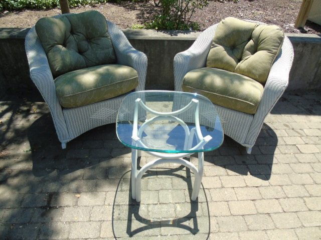 Lot of 3 Outdoor Furniture Pieces: A lot of three outdoor furniture pieces. Two woven wicker rocking chairs with one loose seat and one loose button back cushion. 33" W x 38" D x 30" H. One glass top end table with painted white metal