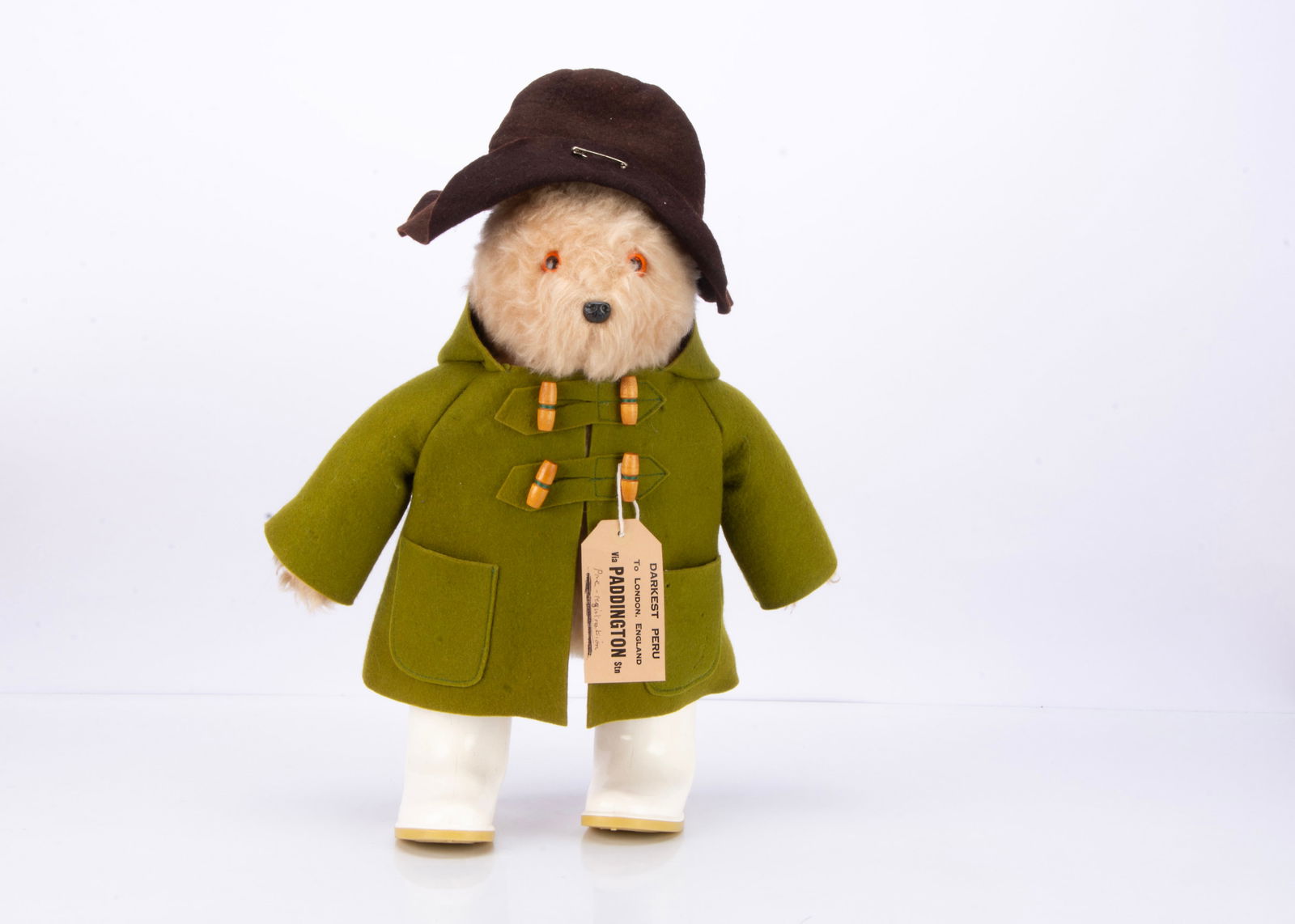 A Gabrielle Deigns Paddington Bear,: A Gabrielle Deigns Paddington Bear, with green duffle coat, dark drown hat, white Dunlop wellies, luggage label with hand written note 'Pre -registration' ó18in. (46cm.) high (some small nibble t