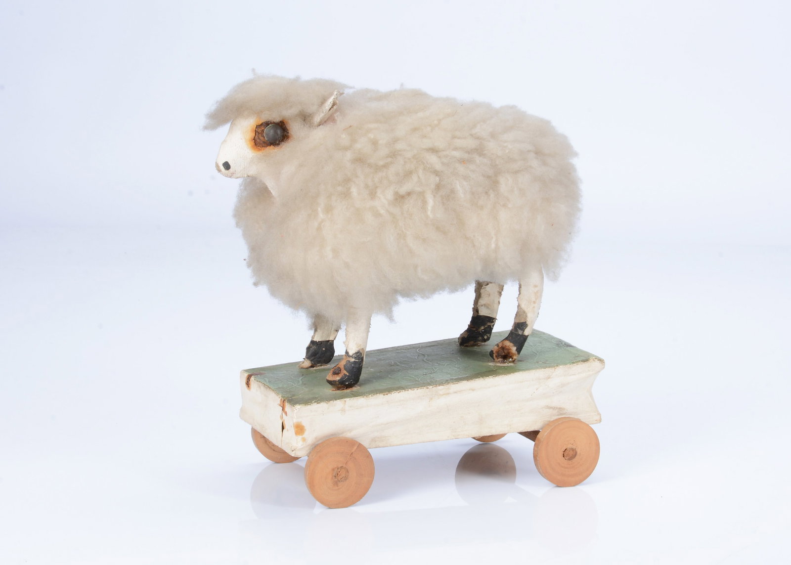 An 19th century German papier-mâché sheep on wheels squeak toy,: An 19th century German papier-mâché sheep on wheels squeak toy, with sheepskin and kid covered face, ears and legs, on wooden base covered in green foliage embossed paper, a kid squeaker bel