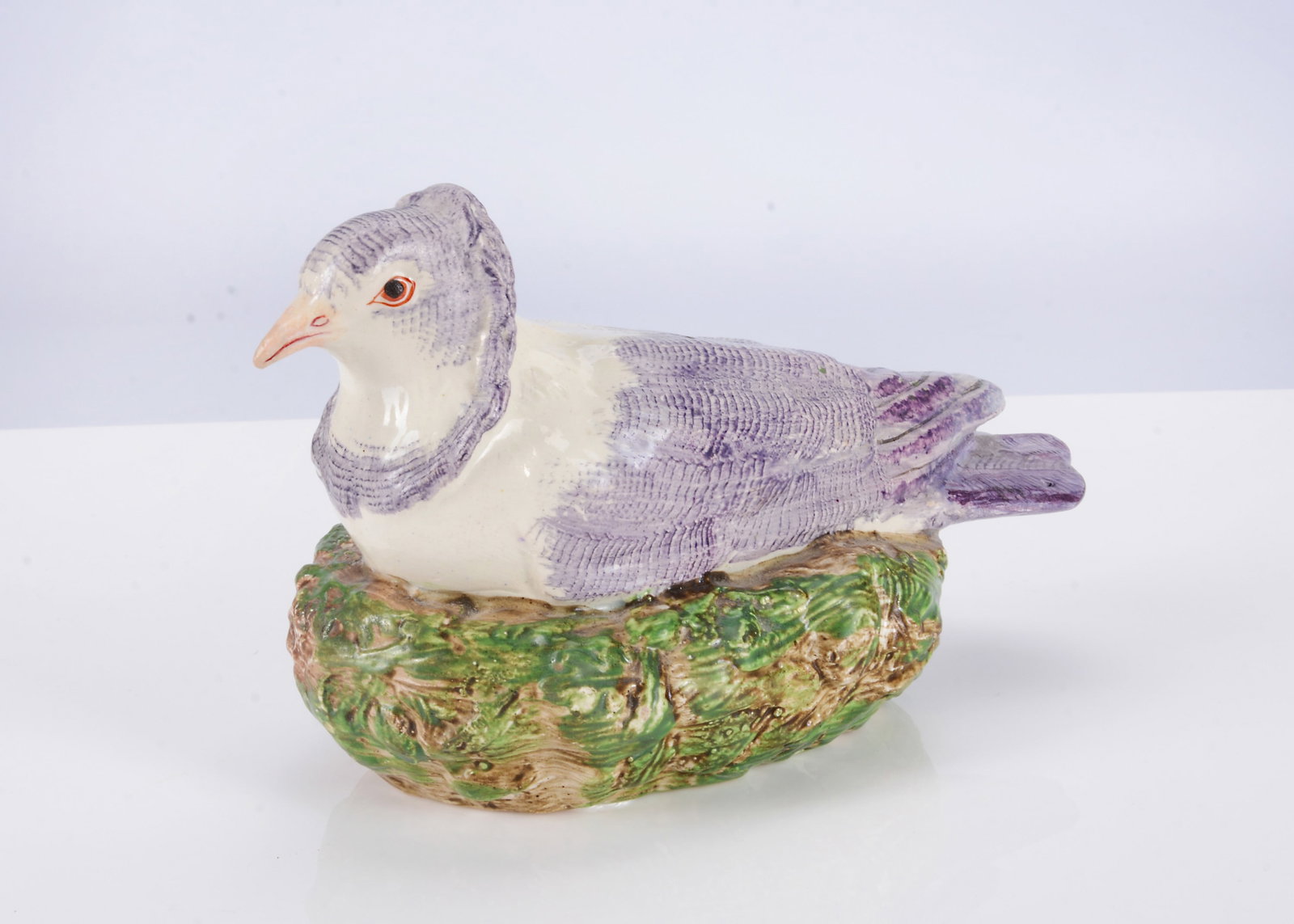 A Staffordshire pearlware pigeon, circa 1800,: A Staffordshire pearlware pigeon, circa 1800, possibly a Nun pigeon, painted with purple feathers, seated on a nest —6 3/4in. (17cm.) long (well restored beak and tail feather) - from Philip Car