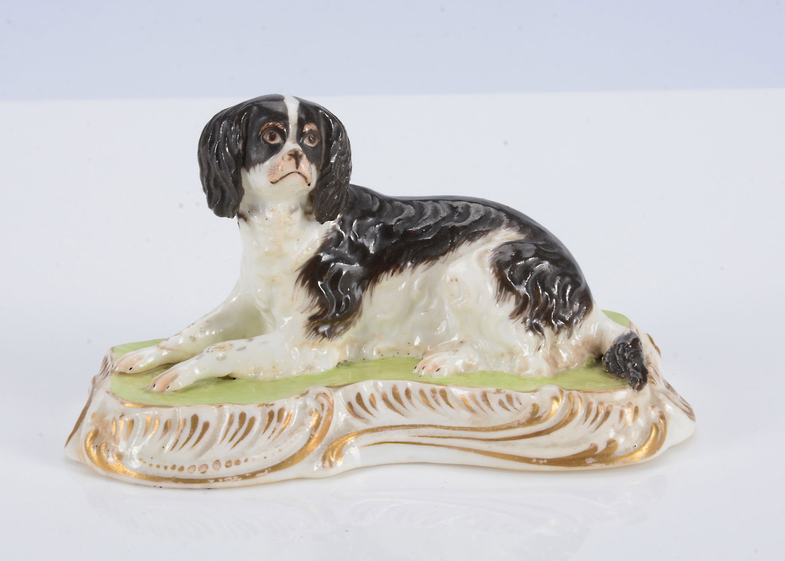 An English porcelain lying spaniel, circa 1835,: An English porcelain lying spaniel, circa 1835, well modelled looking up and to the left, black and white with light brown detail, painted green base with rococo gilt edge —4 1/2in. (11.5cm.) lo