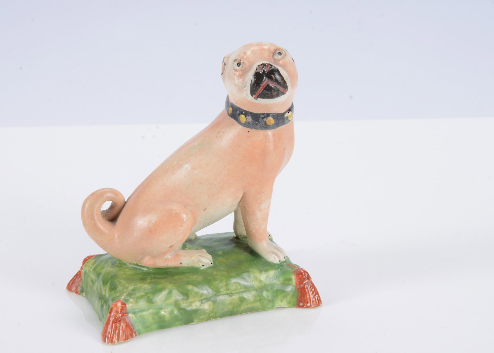 An English pearlware pug dog seated on a cushion, circa 1820,: An English pearlware pug dog seated on a cushion, circa 1820, painted light brown with black collar with yellow studs, head turned to the right, seated on a green painted cushion with dark red tassels