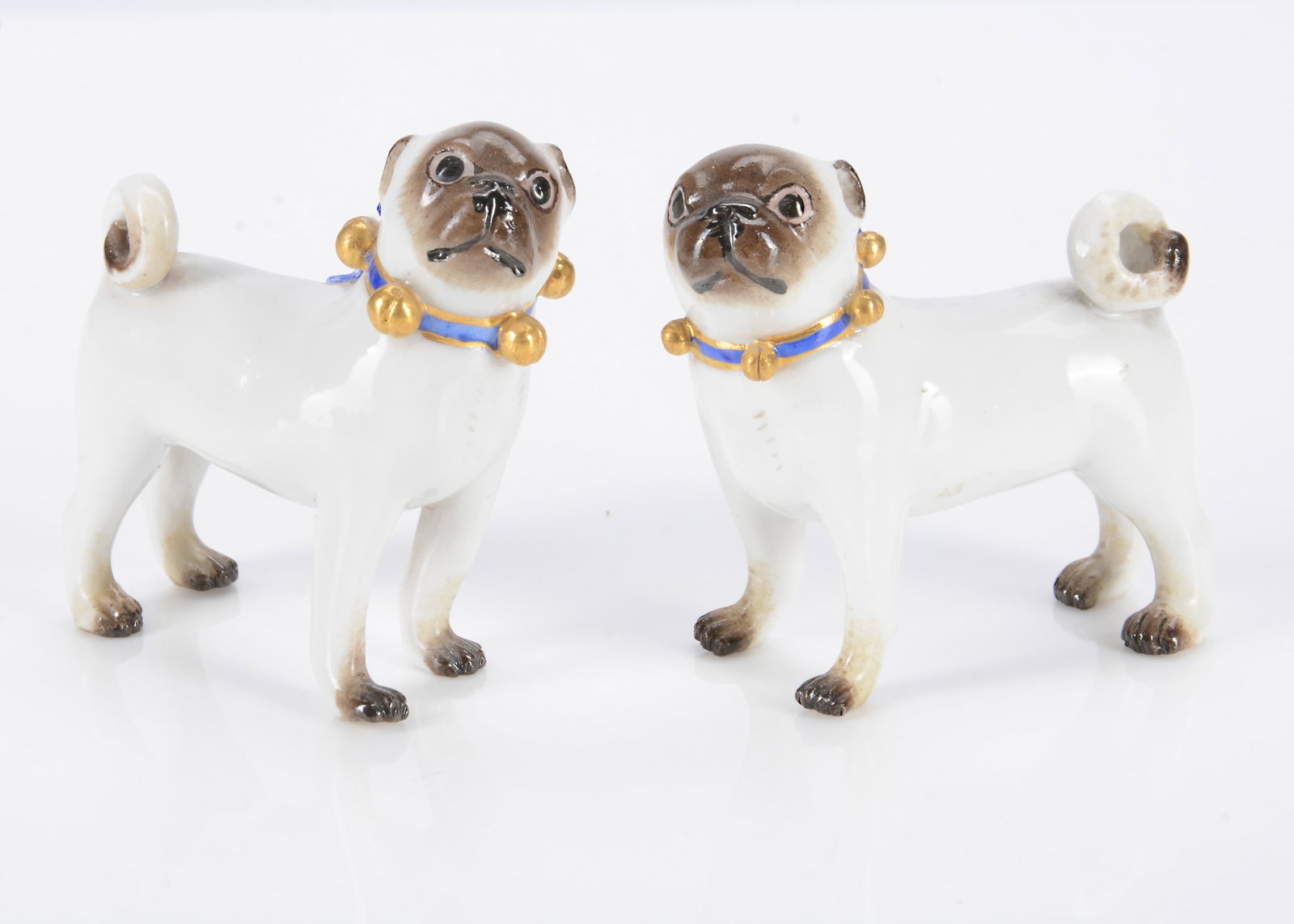 A late 19th century pair of Meissen standing pug dogs,: A late 19th century pair of Meissen standing pug dogs, with brown painted faces and paws, the dog slightly larger with slightly larger blue and gilt ribbon with gilt bells, the bitch similar and sligh