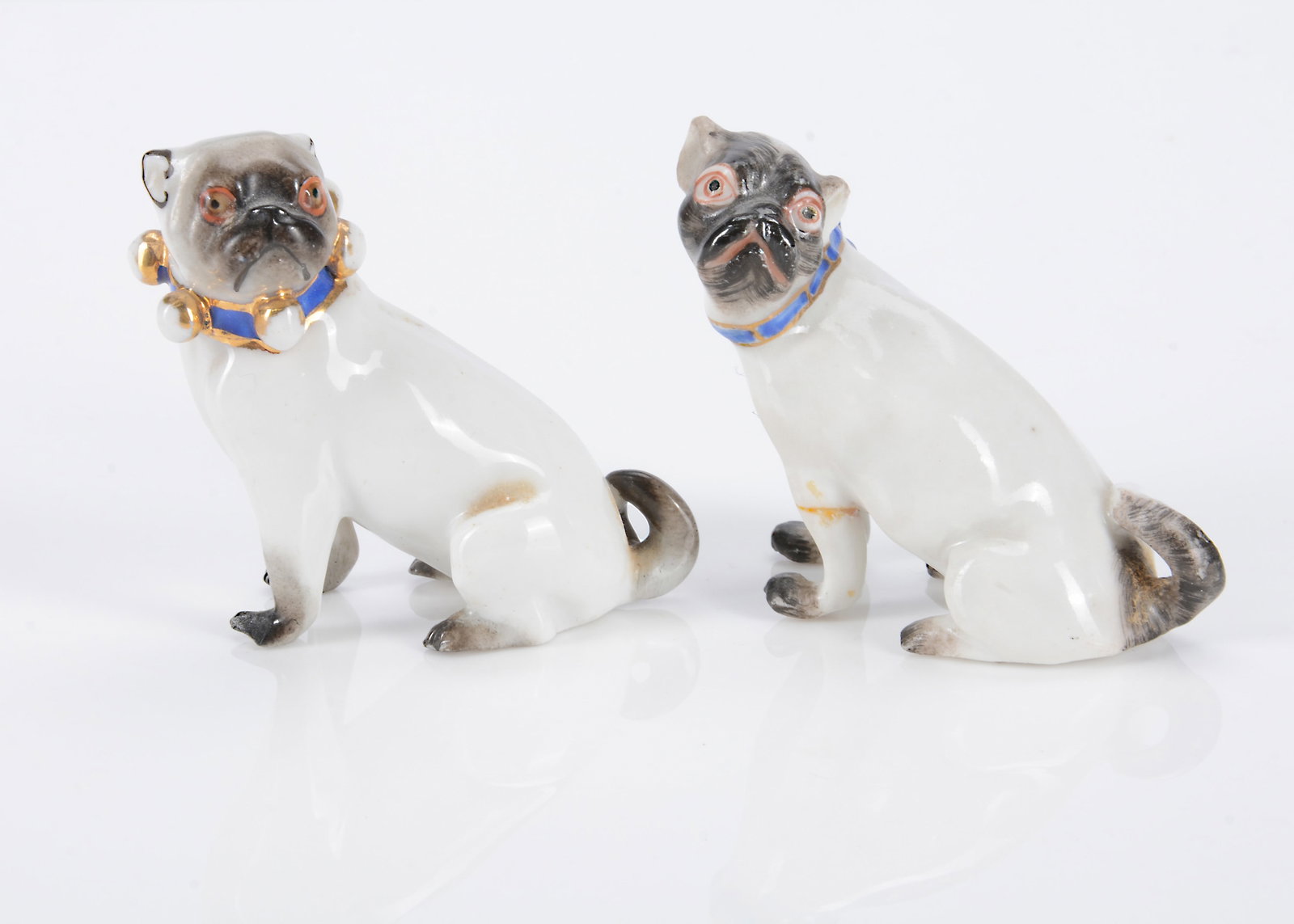 Two German porcelain pug dogs, circa 1900,: Two German porcelain pug dogs, circa 1900, one with blue painted collar with gold bells, seated looking to the left and inscribed 103 —2in. (5cm.) high (bells rubbed); and another seated looking