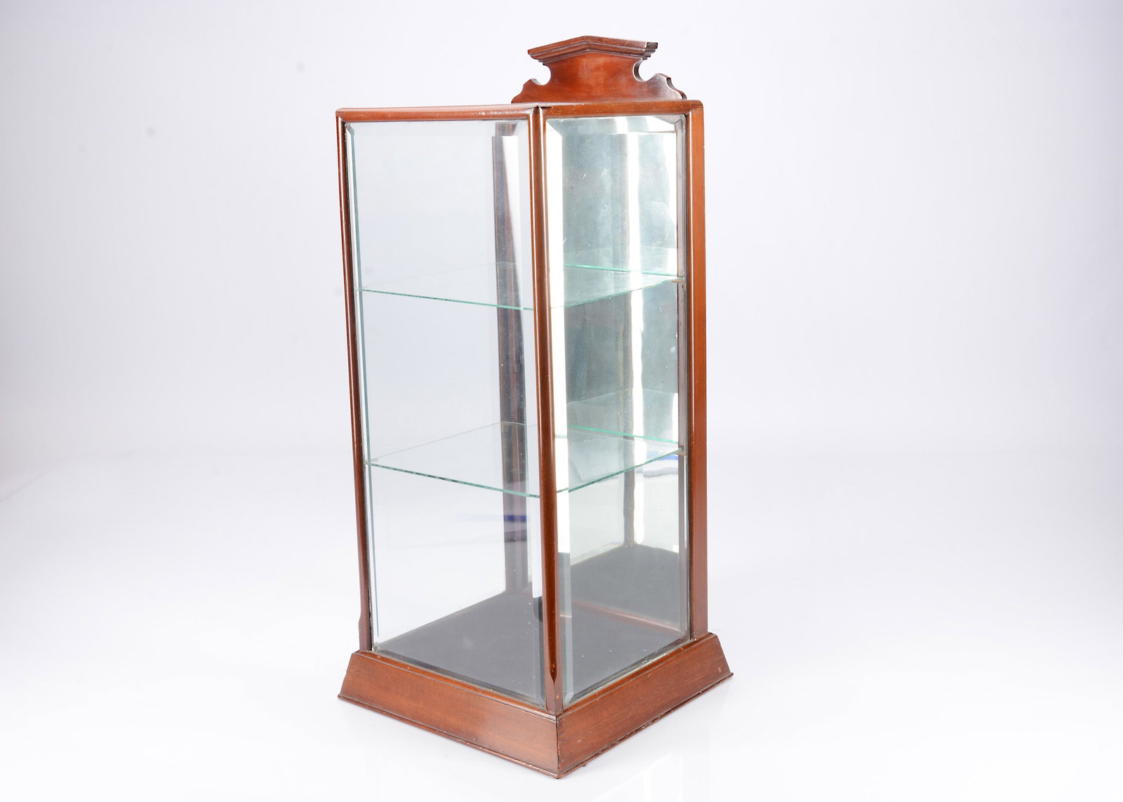 A mahogany counter top display cabinet, circa 1900,: A mahogany counter top display cabinet, circa 1900, with pediment, bevelled glass, rear opening mirrored back and two glass shelves —27in. (68.5cm.) high (one shelf with corner missing)