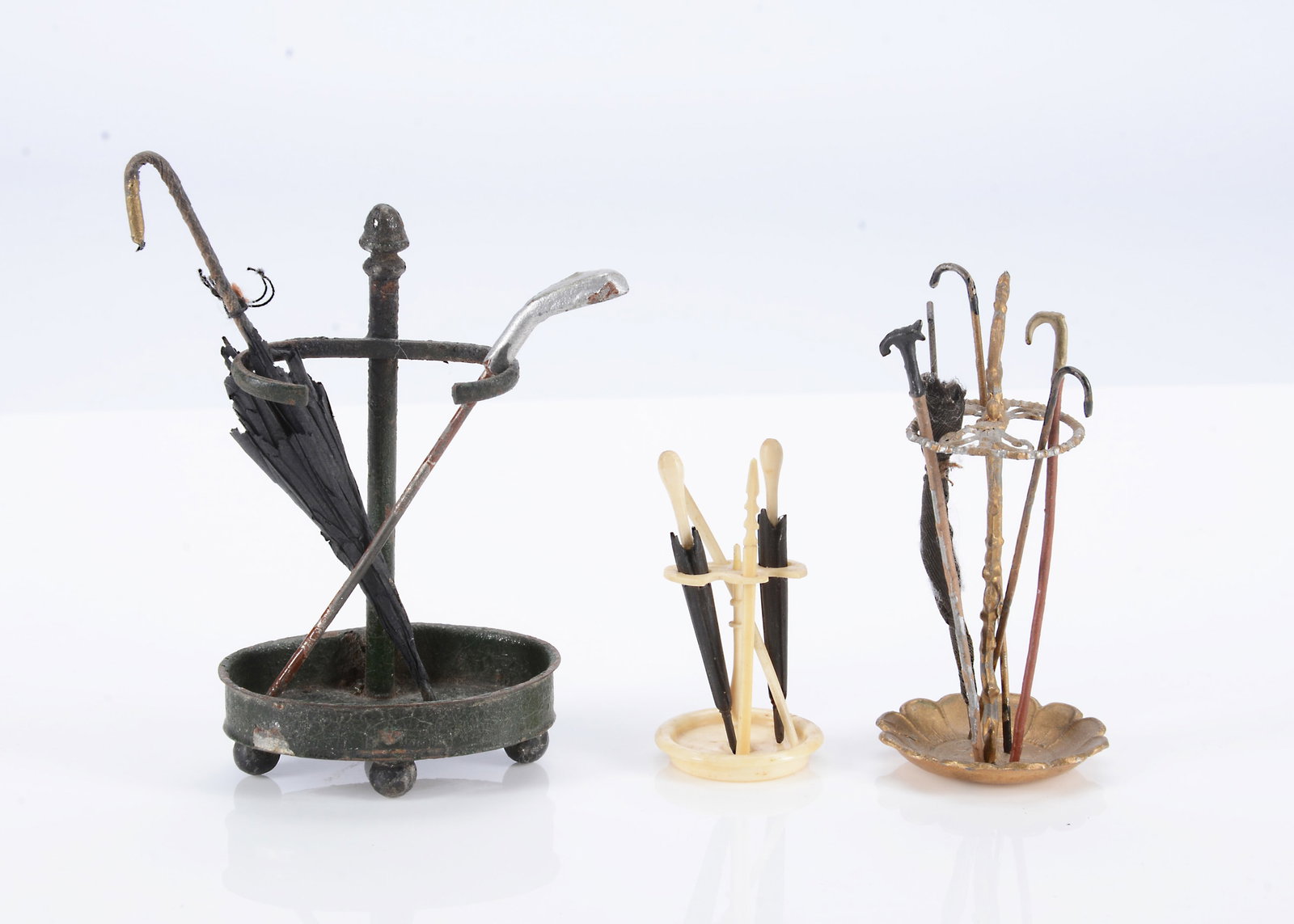 Three 19th century German dolls’ house stick stands,: Three 19th century German dolls’ house stick stands, a green painted tinplate example with umbrella and golf club —3 1/2in. (9cm.) high (some rusting); a gold painted soft metal example; a