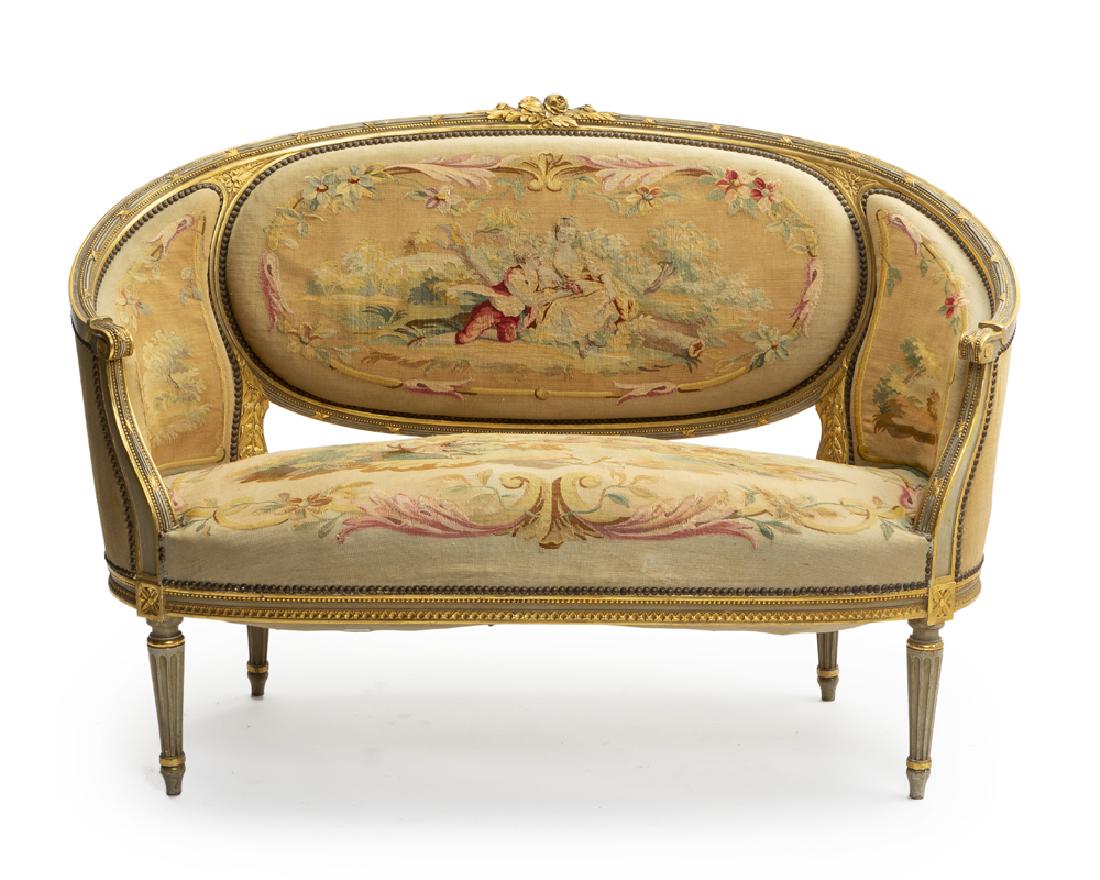 A Louis XVI-style giltwood settee: Late 19th/early 20th century The curved carved and painted giltwood frame enclosing an oval backrest, two solid side rests, and an oblong seat raised on tapering fluted legs, upholstered with a tapest