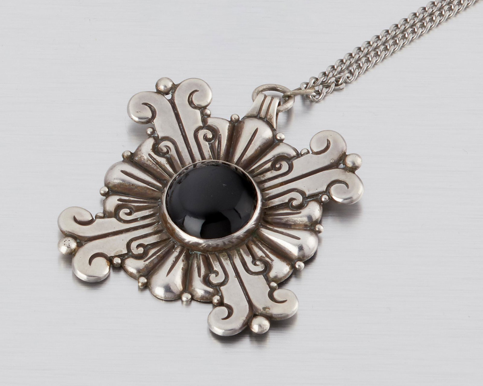 A Hector Aguilar silver and obsidian pendant/brooch: 1940-1945; Taxco, Mexico Stamped for Hector Aguilar; Further stamped: Taxco / 990 The Maltese cross-style pendant/brooch centering a round obsidian cabochon suspended from an attached chain 24" L;