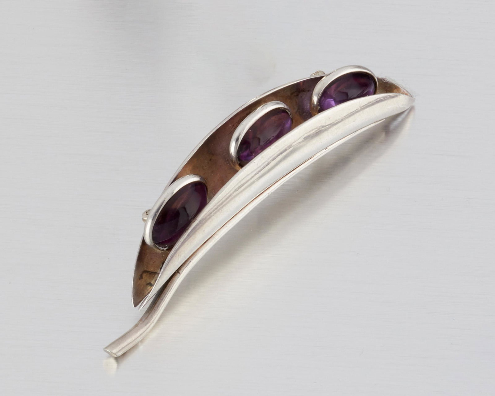 An Antonio Pineda silver and amethyst brooch: 1953-1979; Taxco, Mexico Crown mark for Antonio Pineda; Eagle 17; Further stamped: Sterling / Mexico / Silver / PR571 The pea pod-form brooch enclosing three amethyst cabochons 3.5" W x .75" H 28.0 gr