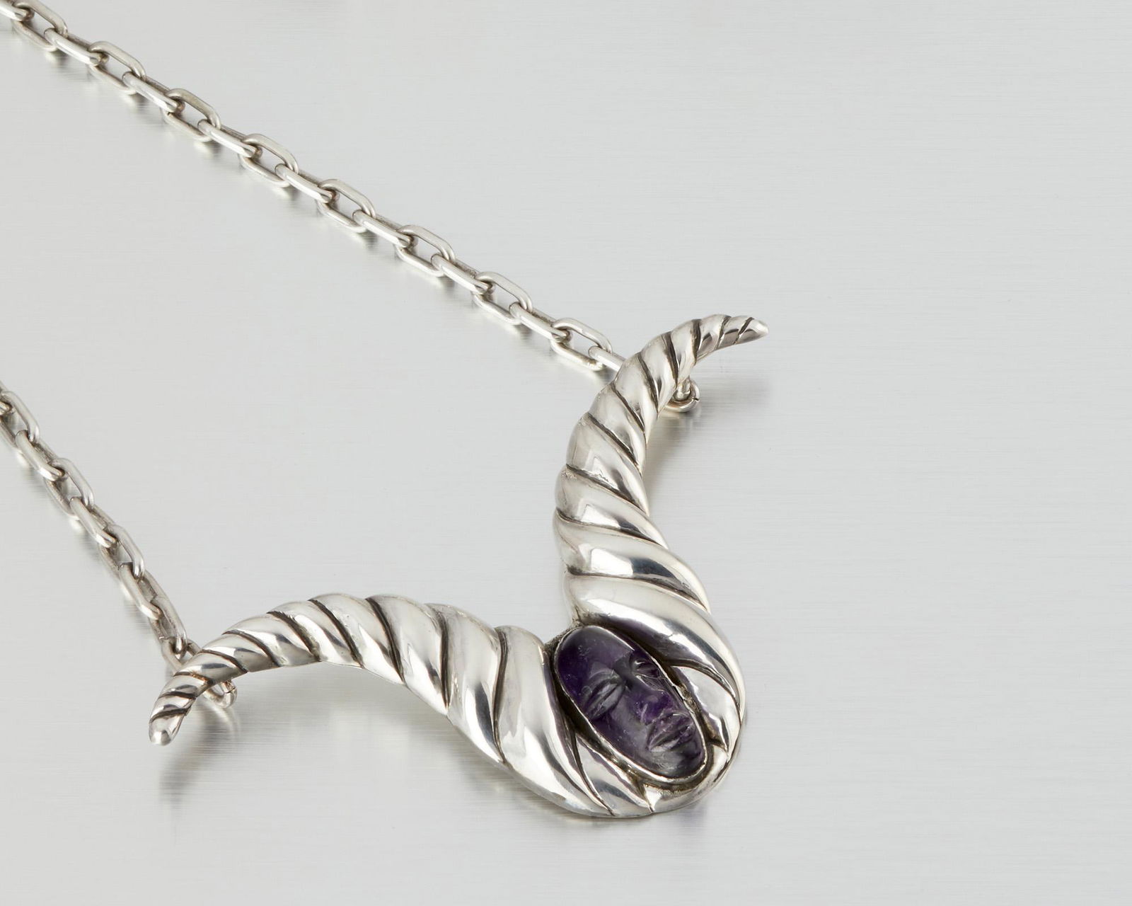 A Hubert Harmon silver and amethyst necklace: 1944-1948; Taxco, Mexico Winged feet for Hubert Harmon Designed with a carved purple stone head with twisted horns attached to a link chain 21" L; Pendant: 2.75" H x 4.75" W 85.0 grams Provenanc