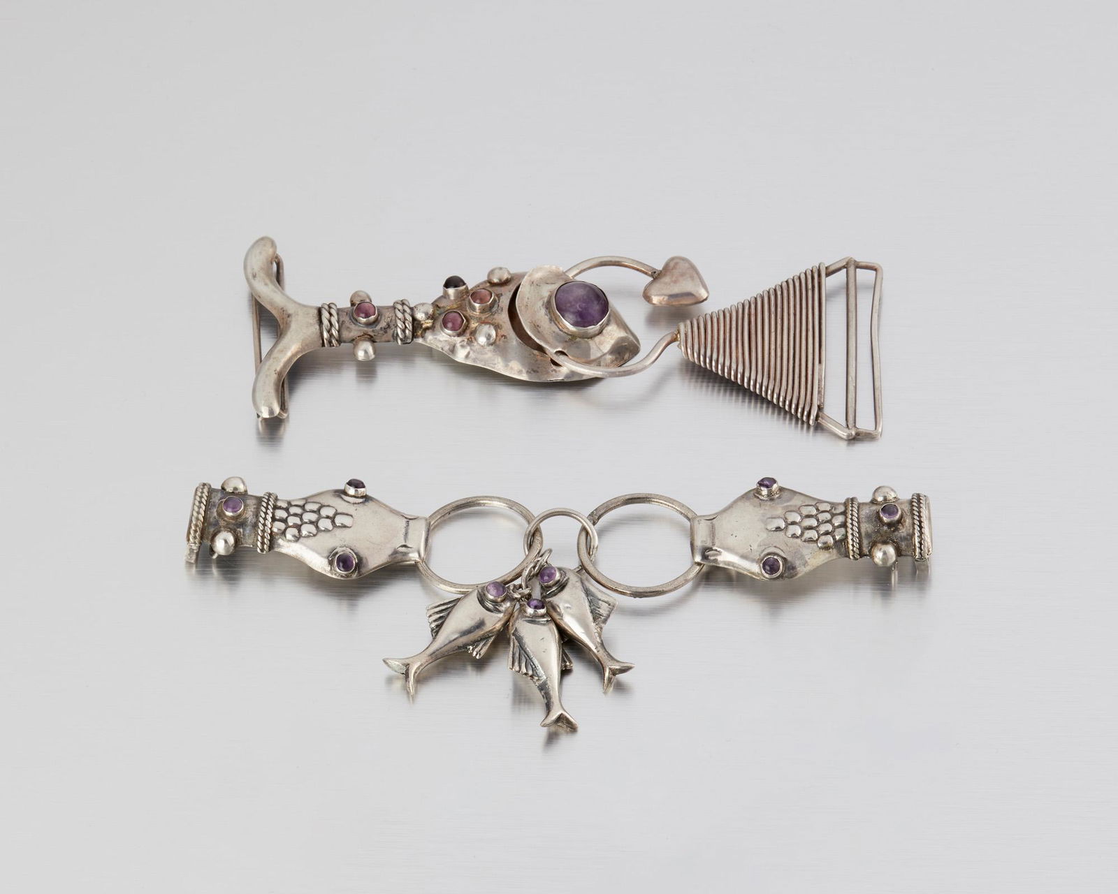 Two Hubert Harmon silver belt buckles: 1944-1948; Taxco, Mexico Winged feet stamp for Hubert Harmon; Further stamped: Hubert Harmon / Sterling / Made in Mexico Comprising a purple stone-set serpent-head belt buckle suspending fish, and a