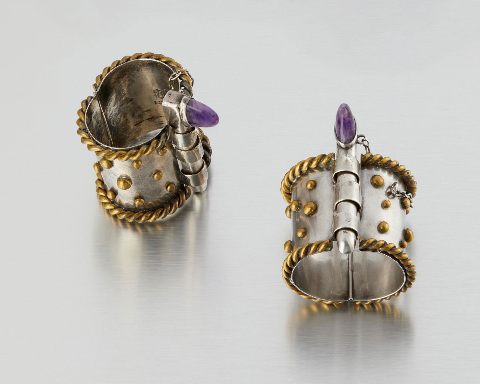 A pair of Hubert Harmon mixed-metal and amethyst bangle: 1942-1948; Taxco, Mexico Winged feet for Hubert Harman; Further stamped: Made in Mexico The silver and brass hinged bracelets with an oversized pin closure set with a cabochon amethyst, 2 pieces Ea