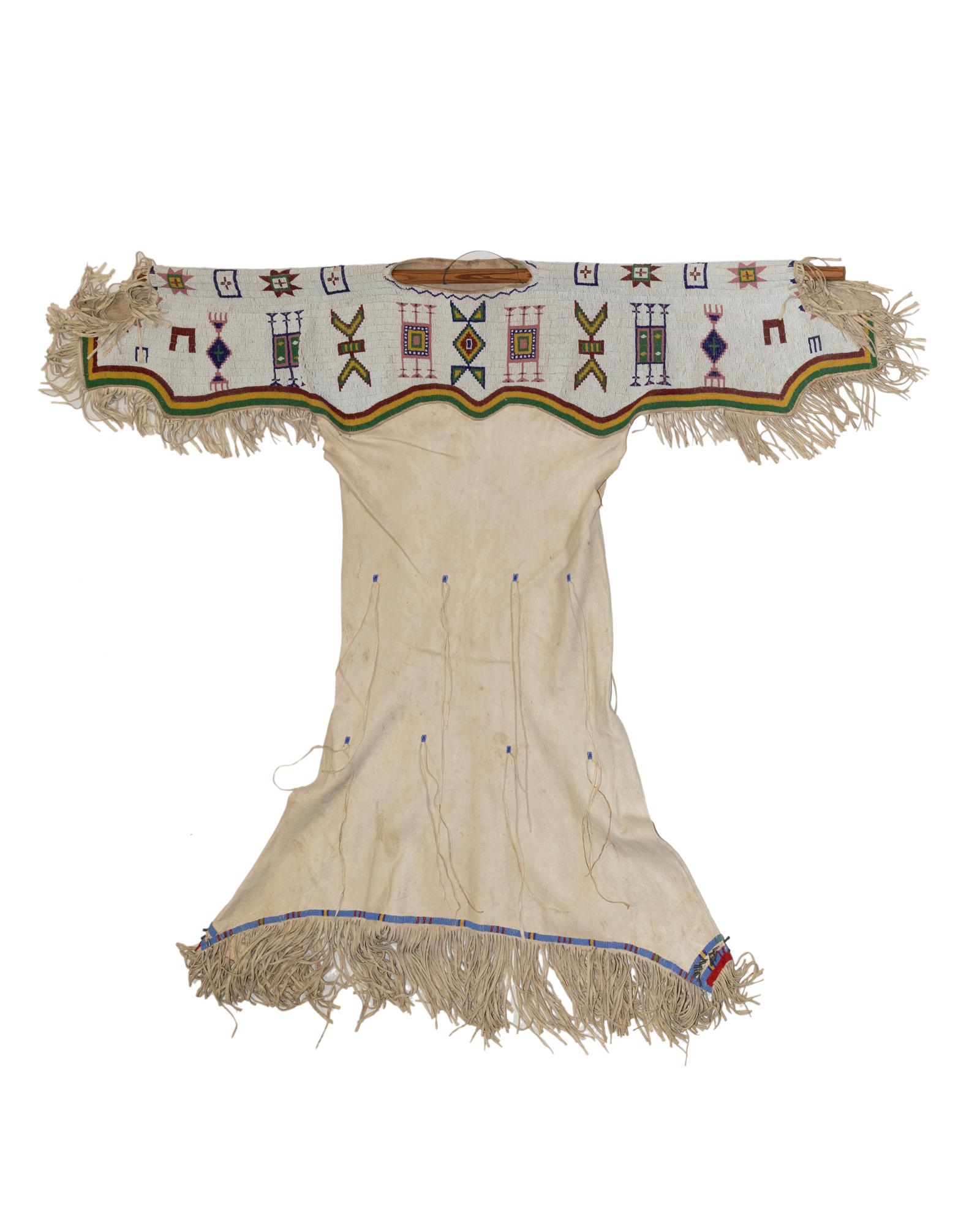 A Northern Plains hide dress: A Northern Plains hide dress Fourth quarter 20th century Designed in buckskin with a polychrome beaded yoke adorned with geometric motifs issuing tin cone fringed edges Without fringe: 51.5" H X 61" W