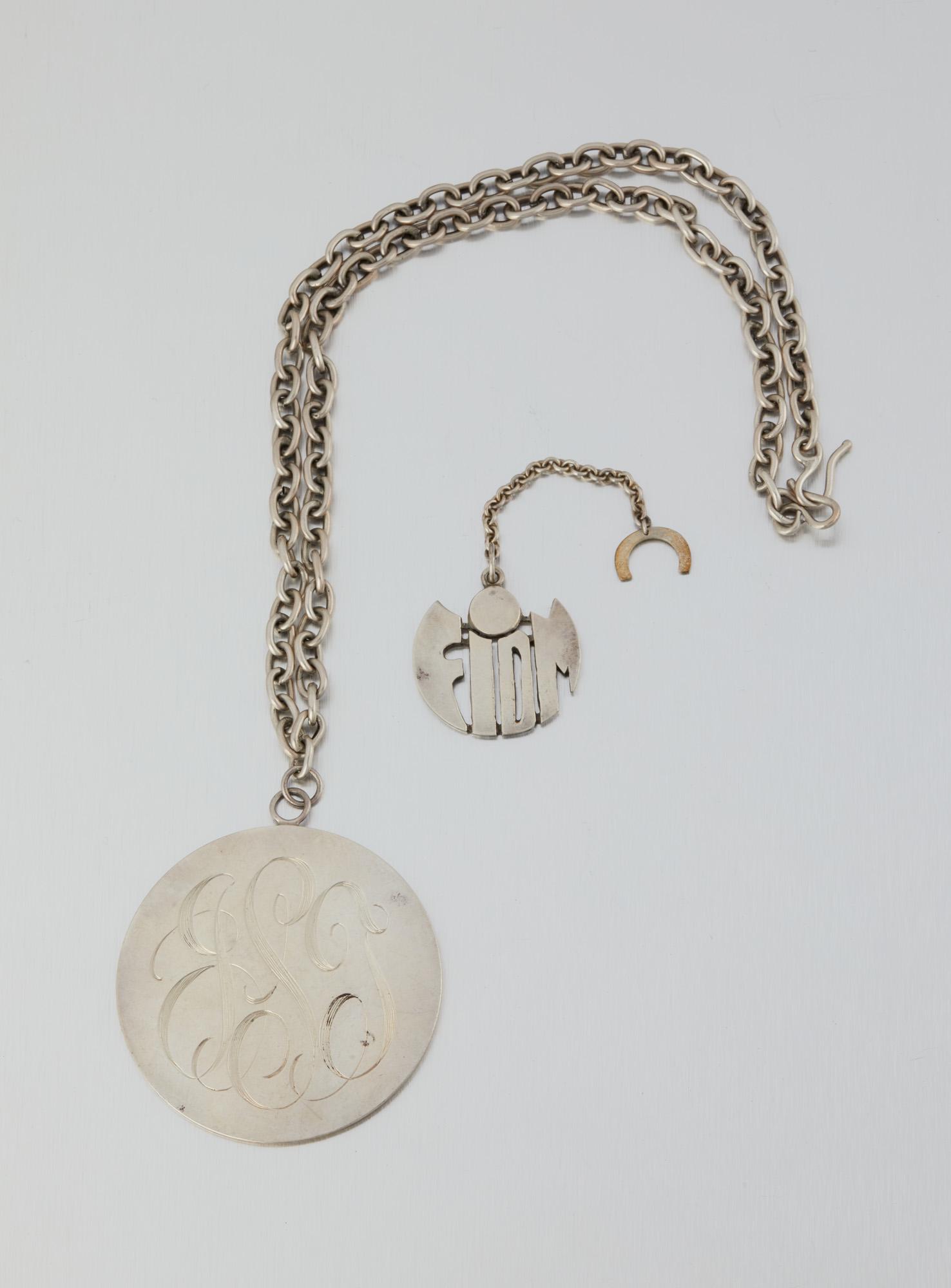 Two Allan Adler jewelry items: Third quarter 20th century; Burbank, CA Each stamped: Allan Adler / Sterling Comprising a FIDM key fob on chain (4.325" L x 1.5" H x 1.5" W); And a large monogramed silver pendant and chain 25" L x