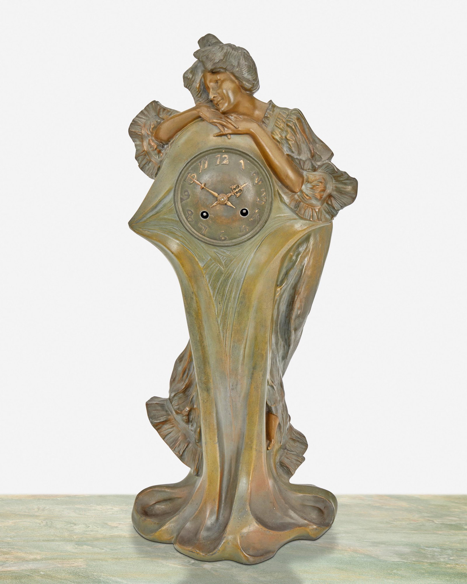 An Art Nouveau patinated metal figural clock: Signed: F. Flora [possibly Francisco Flora] Serial number: 820 The patinated metal clock with low relief gilt Arabic numeral hour markers and two train movement set in a tapered base with a standing f