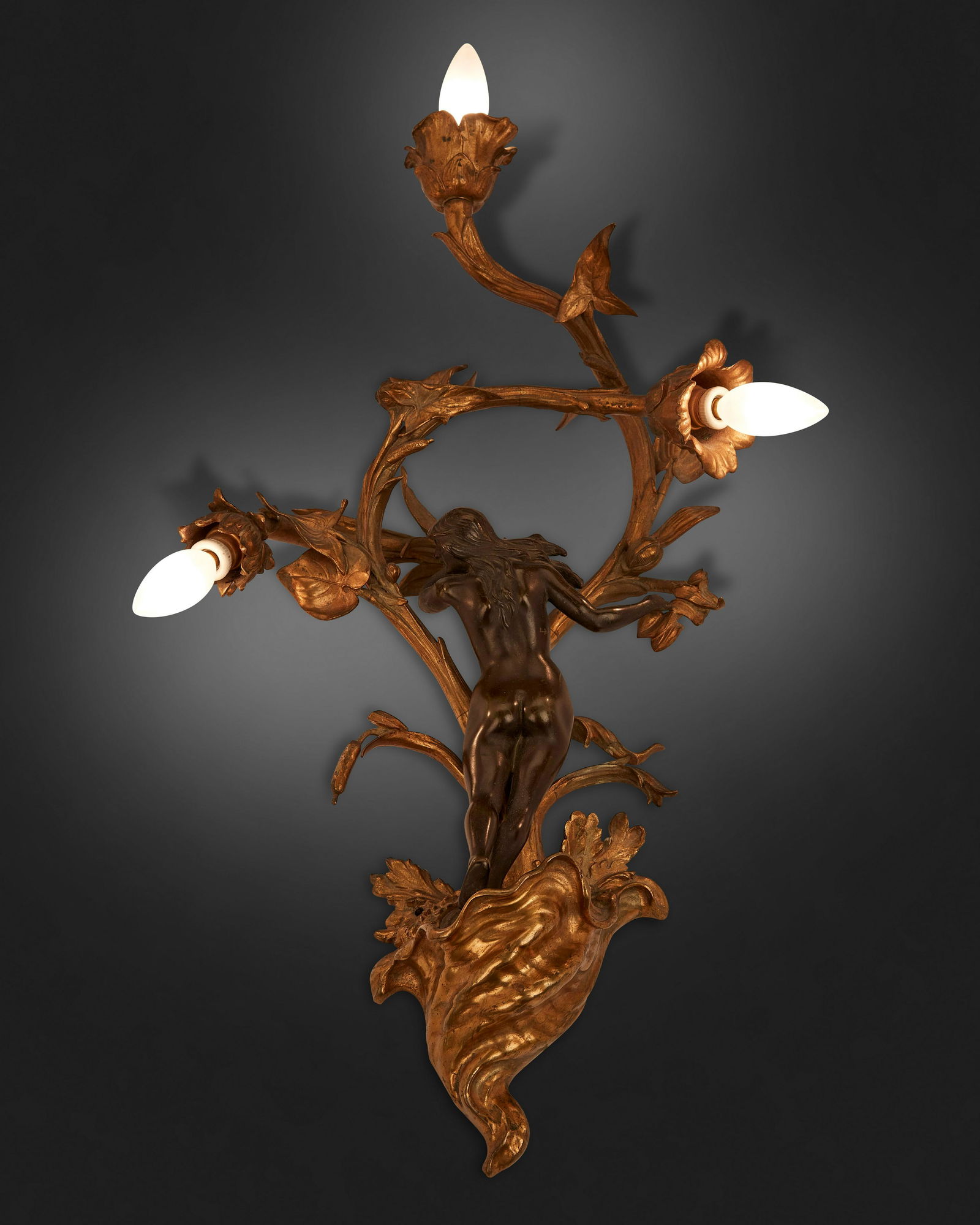 A French Art Nouveau patinated and gilt-bronze wall: First-quarter 20th Century The patinated and gilt-bronze figural wall sconce with female figure standing atop a shell issuing three foliate arms, each terminating in a floral-style capital with light